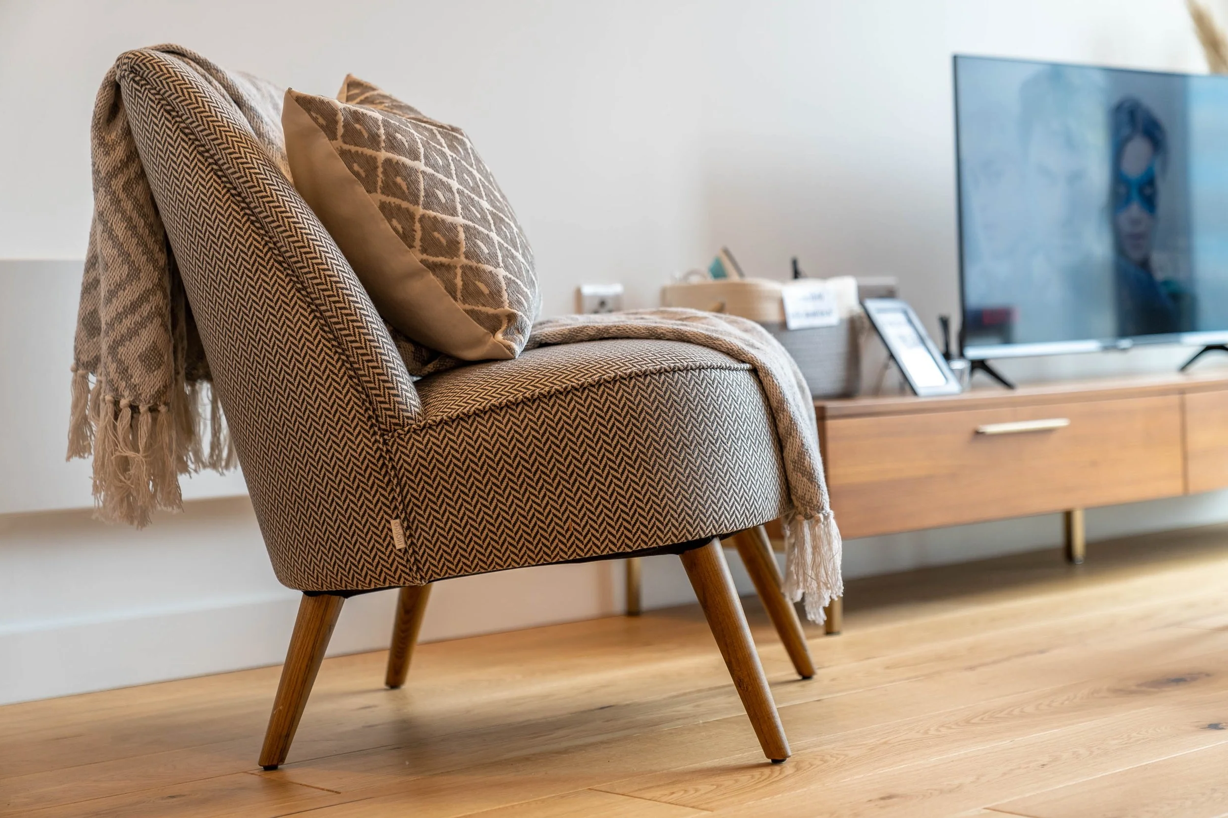 Close-up of a patterned upholstered chair with a beige and brown throw and two decorative cushions, in a modern living room with wooden floors and a media console with TV.