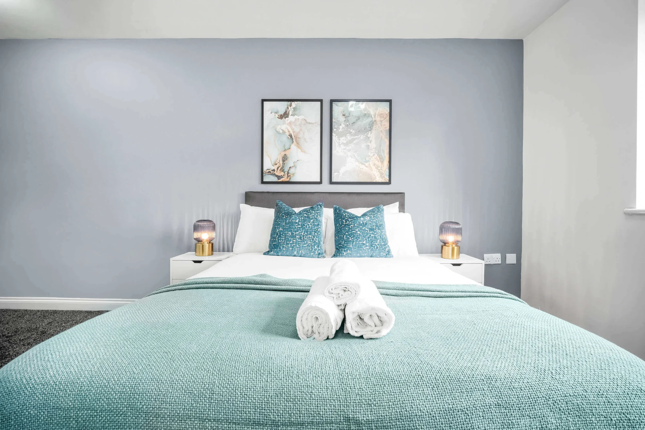 A neatly made bedroom with a bed covered in a teal blanket, two rolled white towels on the bed, two teal throw pillows, a white nightstand on each side with small lamps, and two framed abstract art pieces hanging on a light gray wall above the headbo