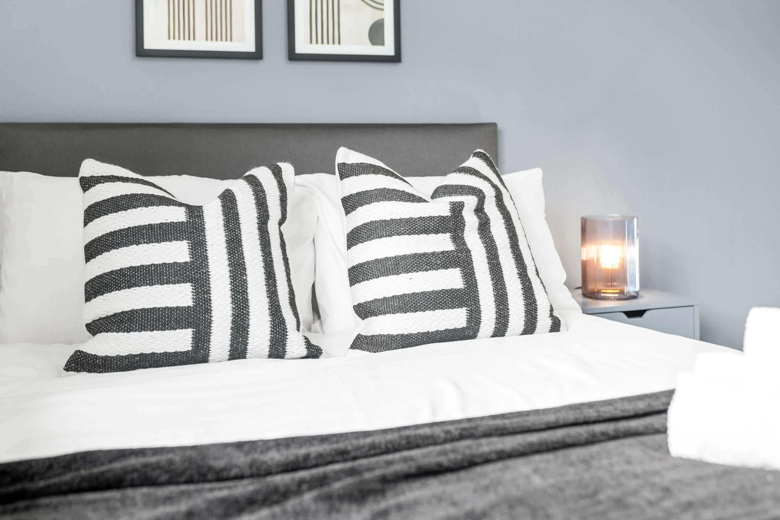 Close-up of a bed with black and white striped pillows, a gray headboard, a white blanket, and a bedside table with a lit candle in a glass holder against a gray wall with two framed abstract art pieces.