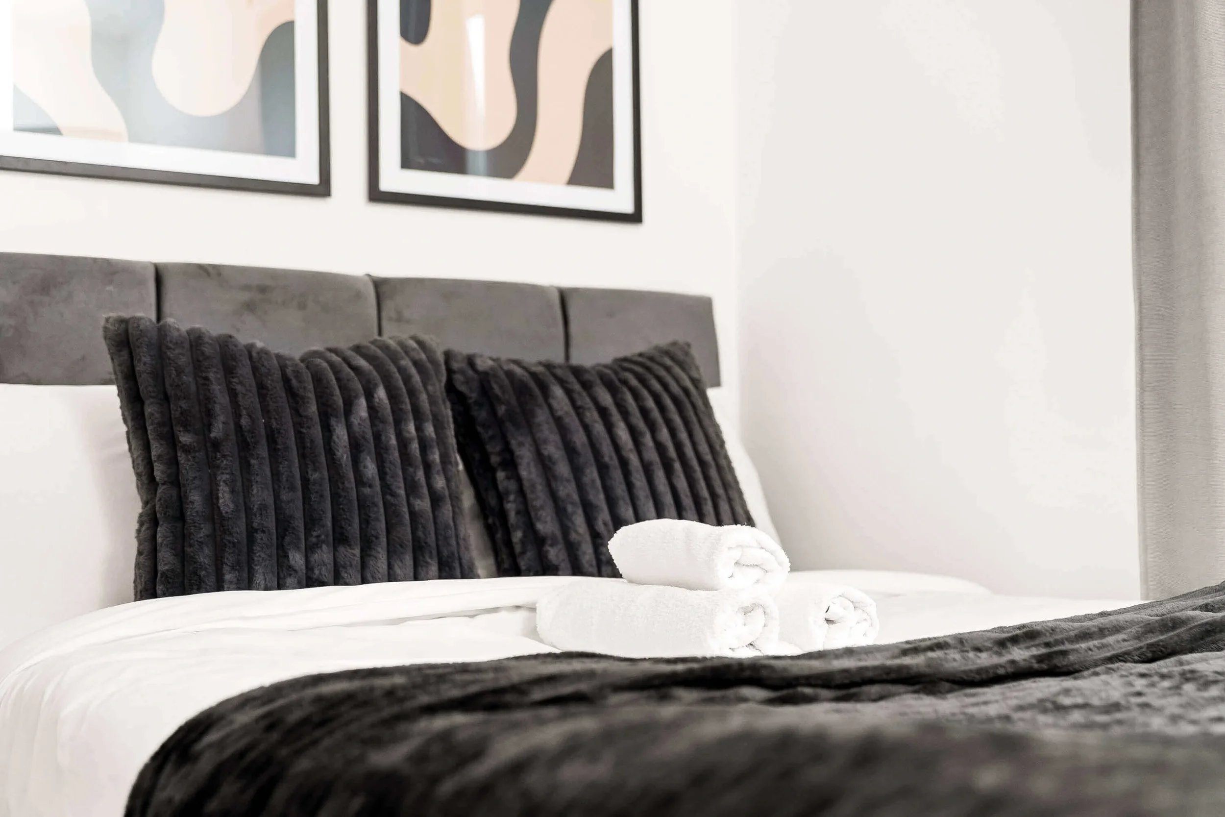 A neatly made bed with white linens, dark gray pillows, and rolled white towels at the foot. Two framed abstract art pieces hang on the wall above the bed.