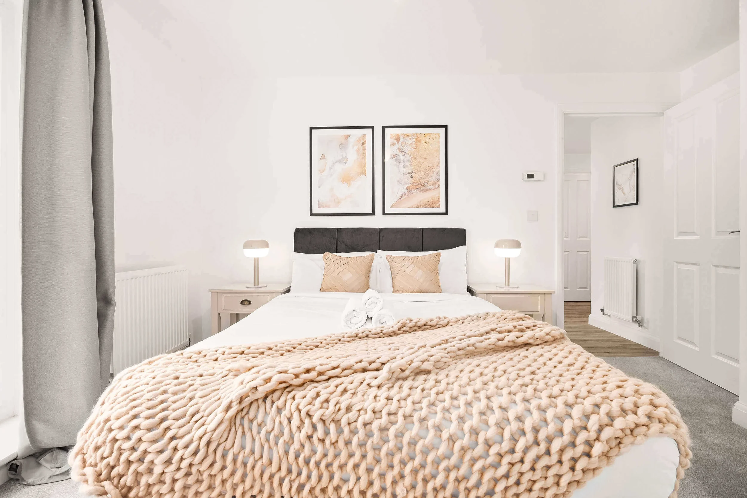 A neatly made bed with a chunky beige blanket, two beige pillows, and white linens in a modern, minimalistic bedroom with white walls, framed abstract art, and bedside lamps.