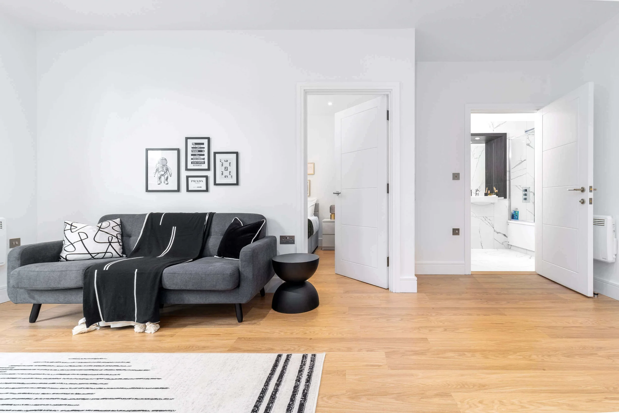 Modern living room with gray sofa decorated with black and white pillows, black side table, and all-white walls with black picture frames. Open door reveals a bathroom with white marble tiles and a sink.