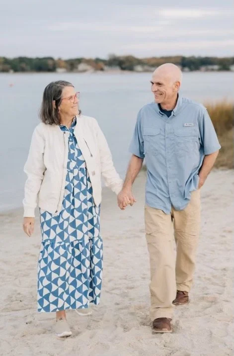An elderly couple walking hand in hand along the beach, smiling at each other.