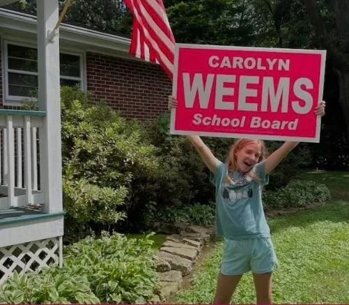 A young girl holding a pink campaign sign that reads 'Carolyn Weems School Board' outside a house with an American flag, surrounded by green plants and grass.