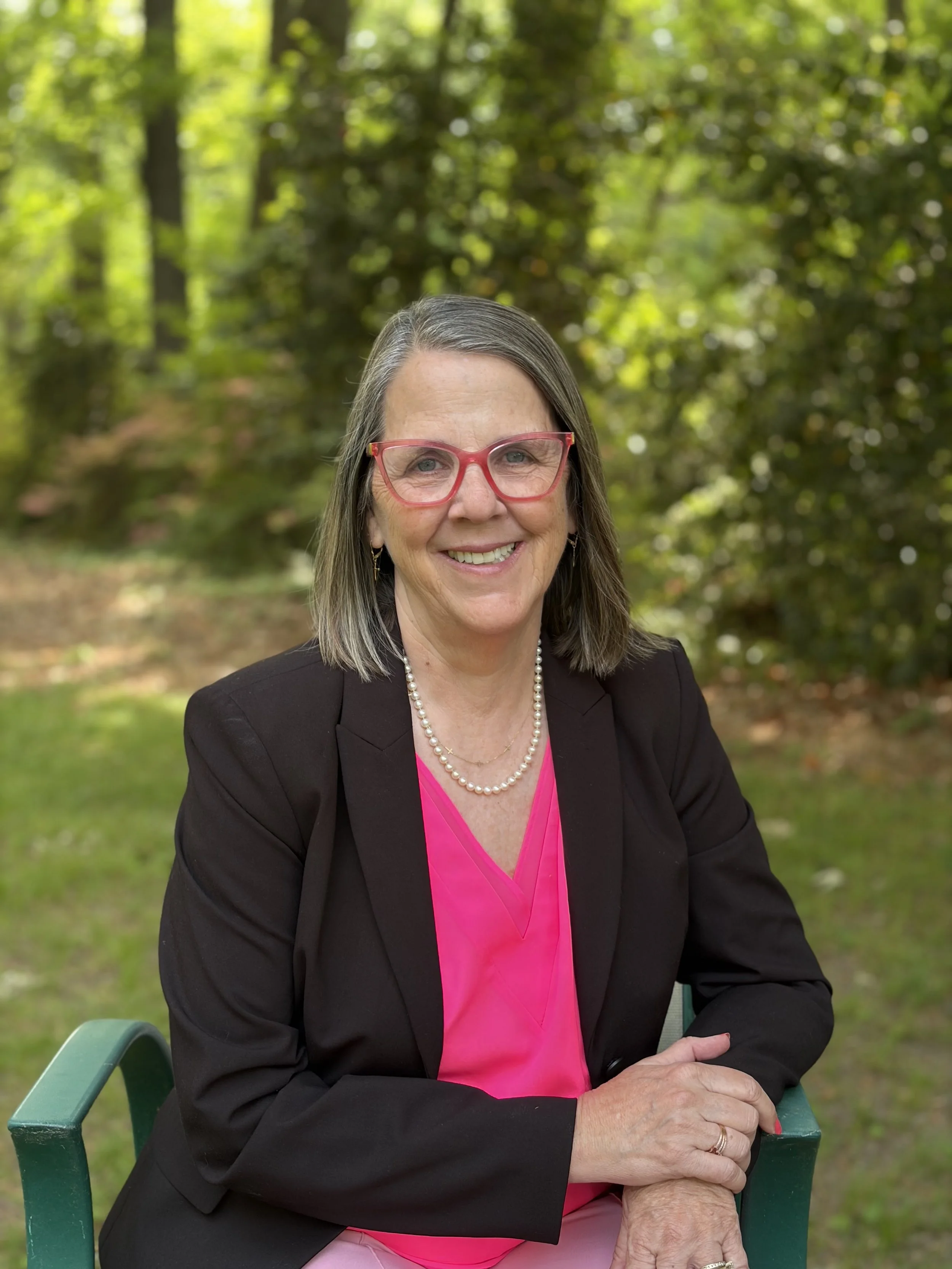 A smiling woman with gray hair, pink glasses, wearing a black blazer, pearl necklace, and pink blouse, sitting outdoors on a green chair in a park with green trees in the background.