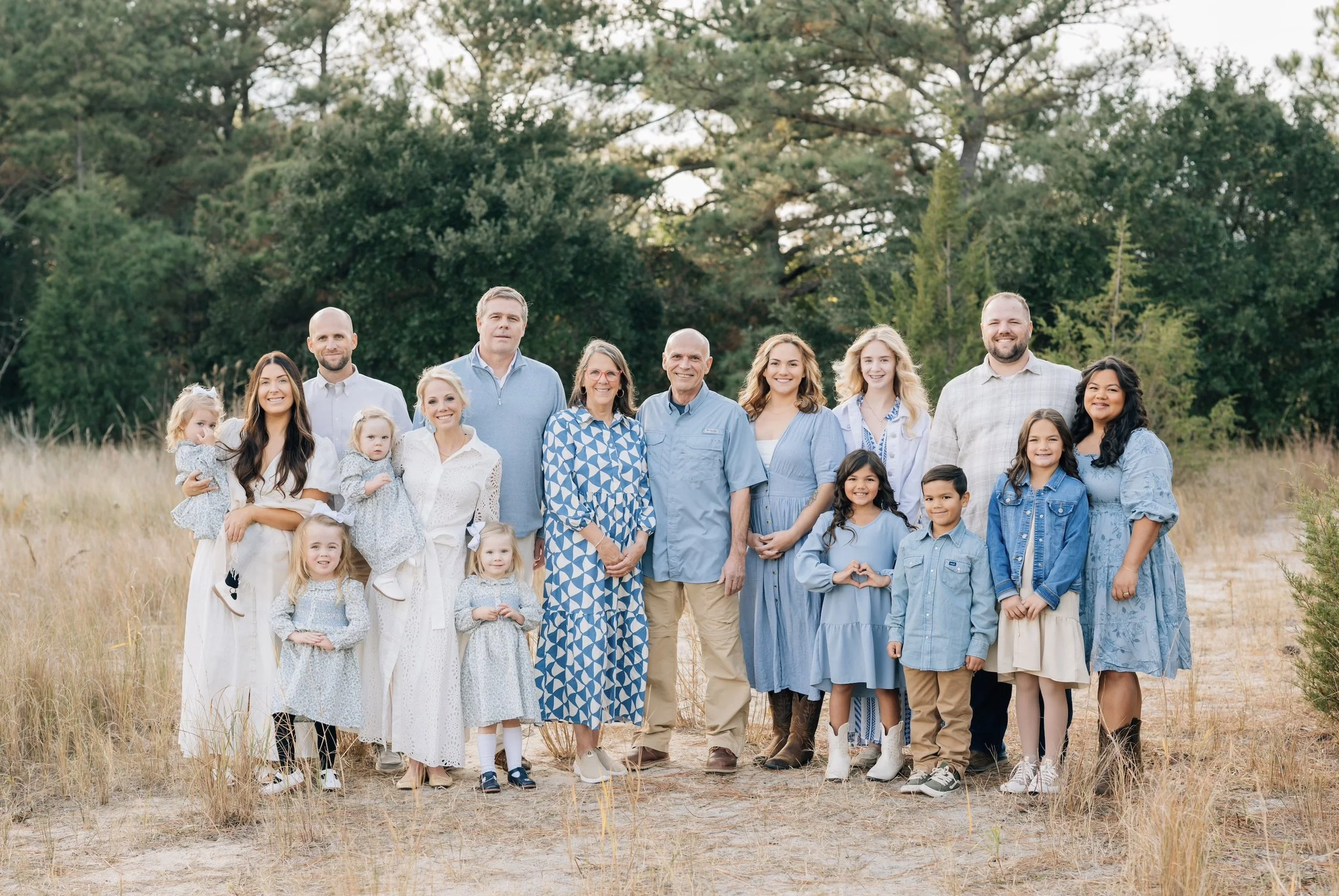 Family group standing outdoors in a grassy area with trees in the background, smiling and posing for a photo.