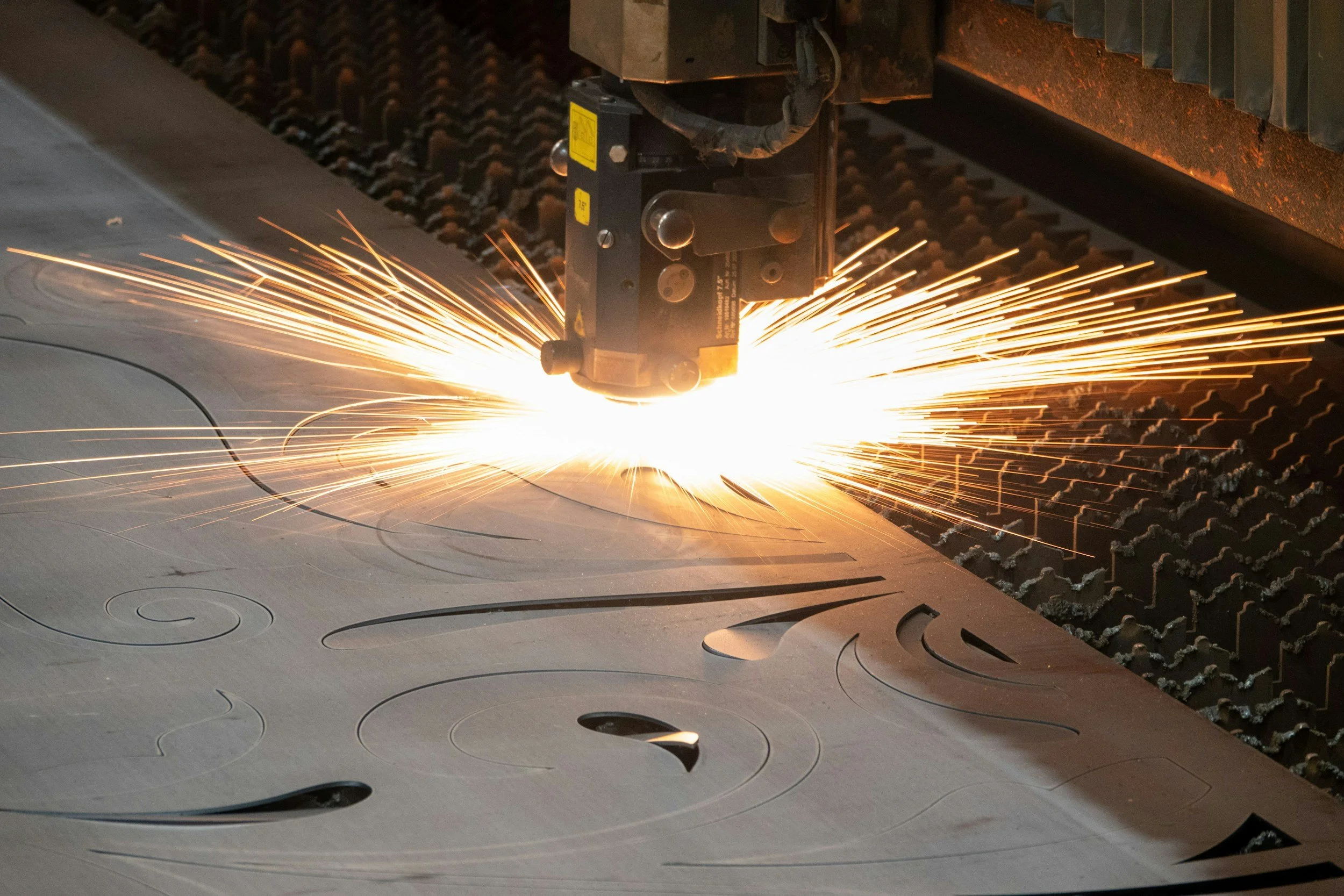 A laser cutter machine engraving and cutting intricate circular patterns into a metal sheet, with sparks flying from the cutting area.