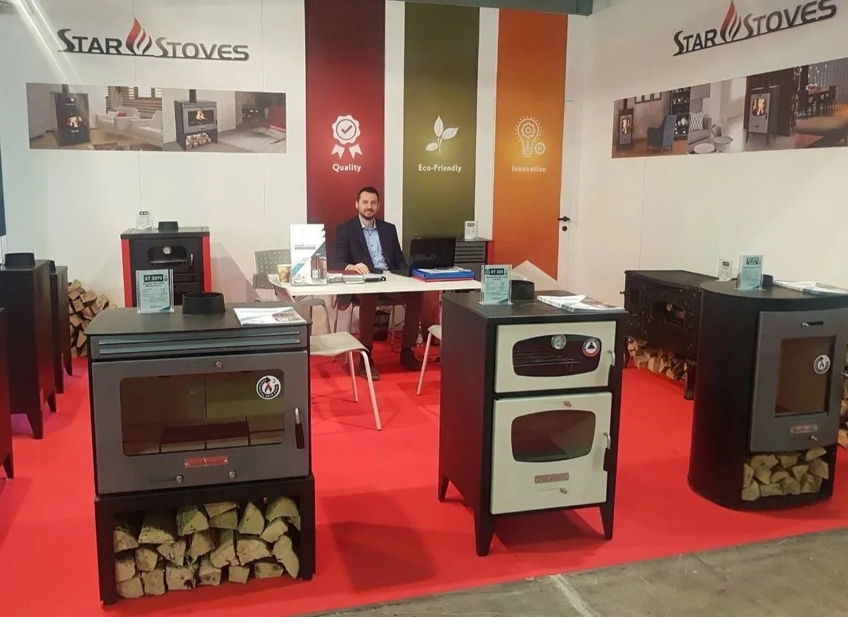 Exhibition booth for Star Stoves displaying various stove models with wood storage below, a man sitting at a desk, and banners highlighting quality, eco-friendliness, and innovation.