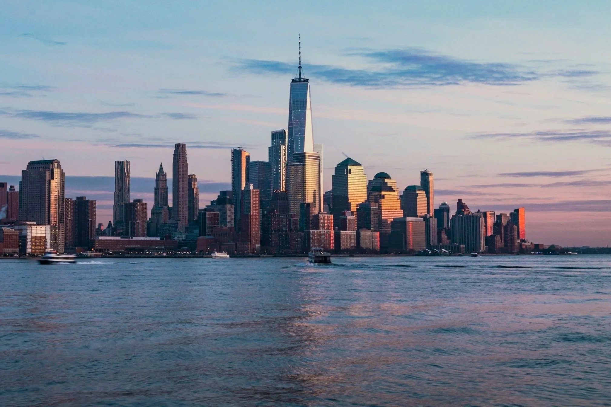 View of New York City skyline at sunset with high-rise buildings and One World Trade Center visible from a waterfront, and boats on the water.