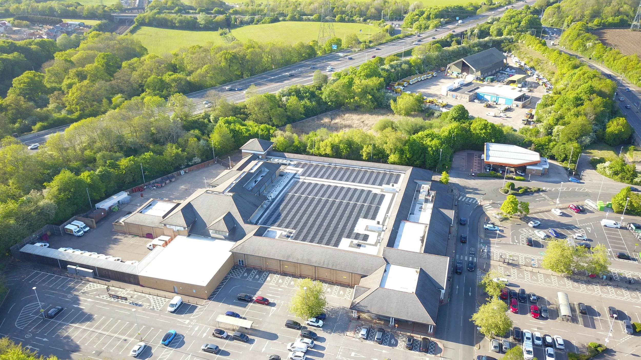 An aerial view of a commercial building with a parking lot, surrounded by trees, a highway with moving cars, and a green area with fields and trees in the background.