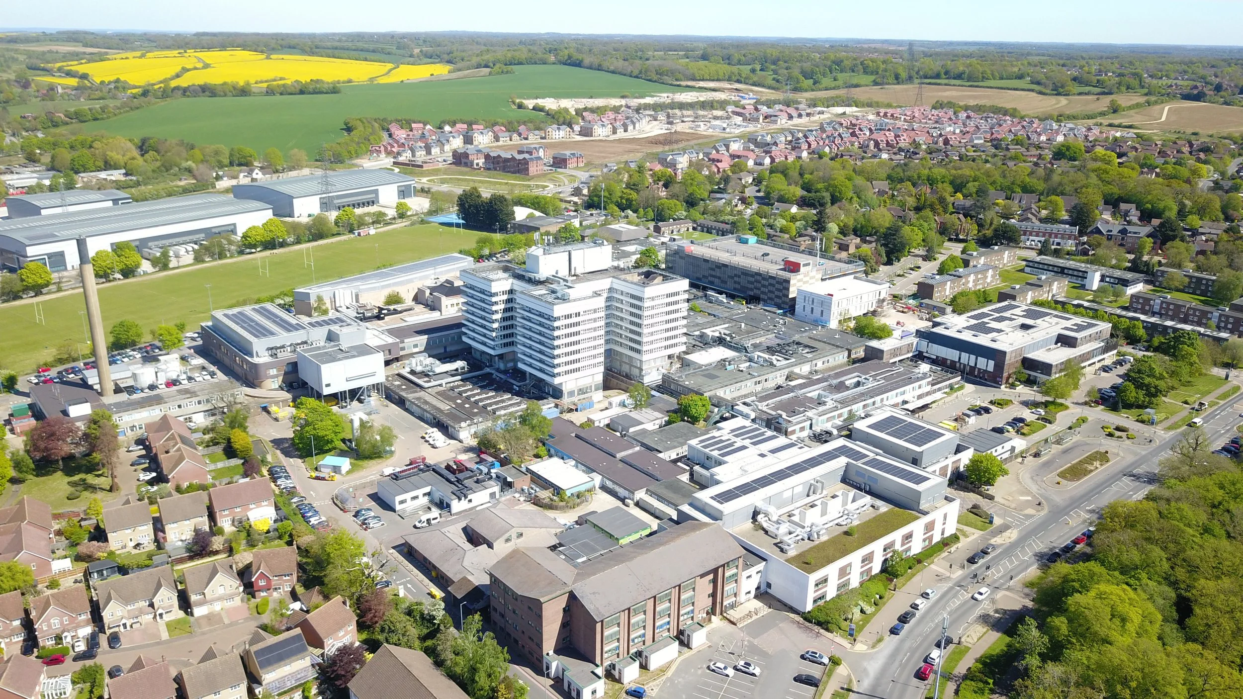 Aerial view of a suburban area with a mix of modern and traditional buildings, green fields, and tree-lined streets on a clear day.