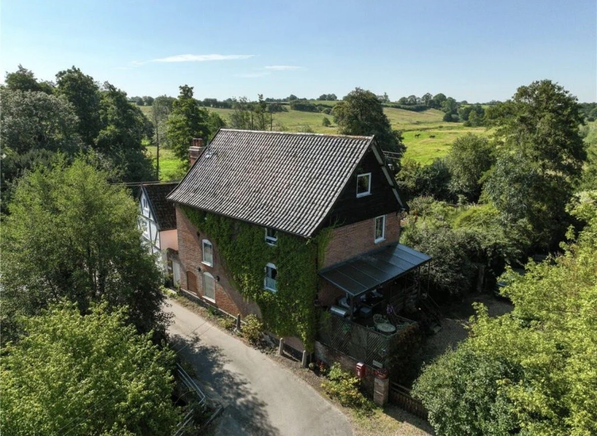 A rural house with a tiled roof, brick walls, and ivy growing up the side, surrounded by trees and green fields, under a blue sky.