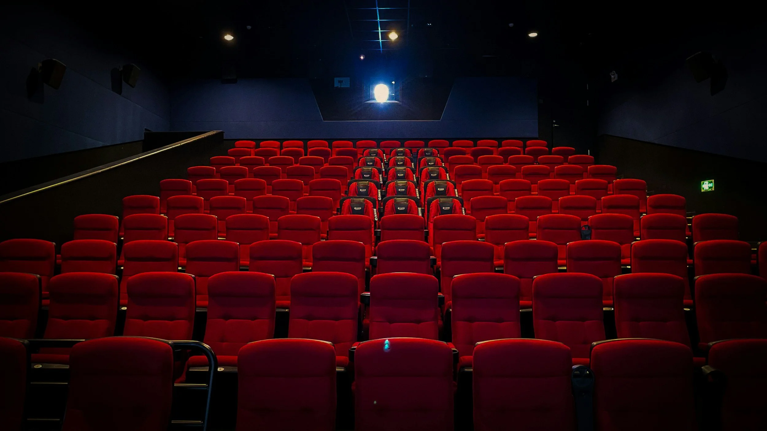 Une salle de cinéma avec des sièges rouges vifs et un écran au fond. L'éclairage est tamisé, avec une lumière bleue provenant de l'écran ou d'une ouverture en haut.