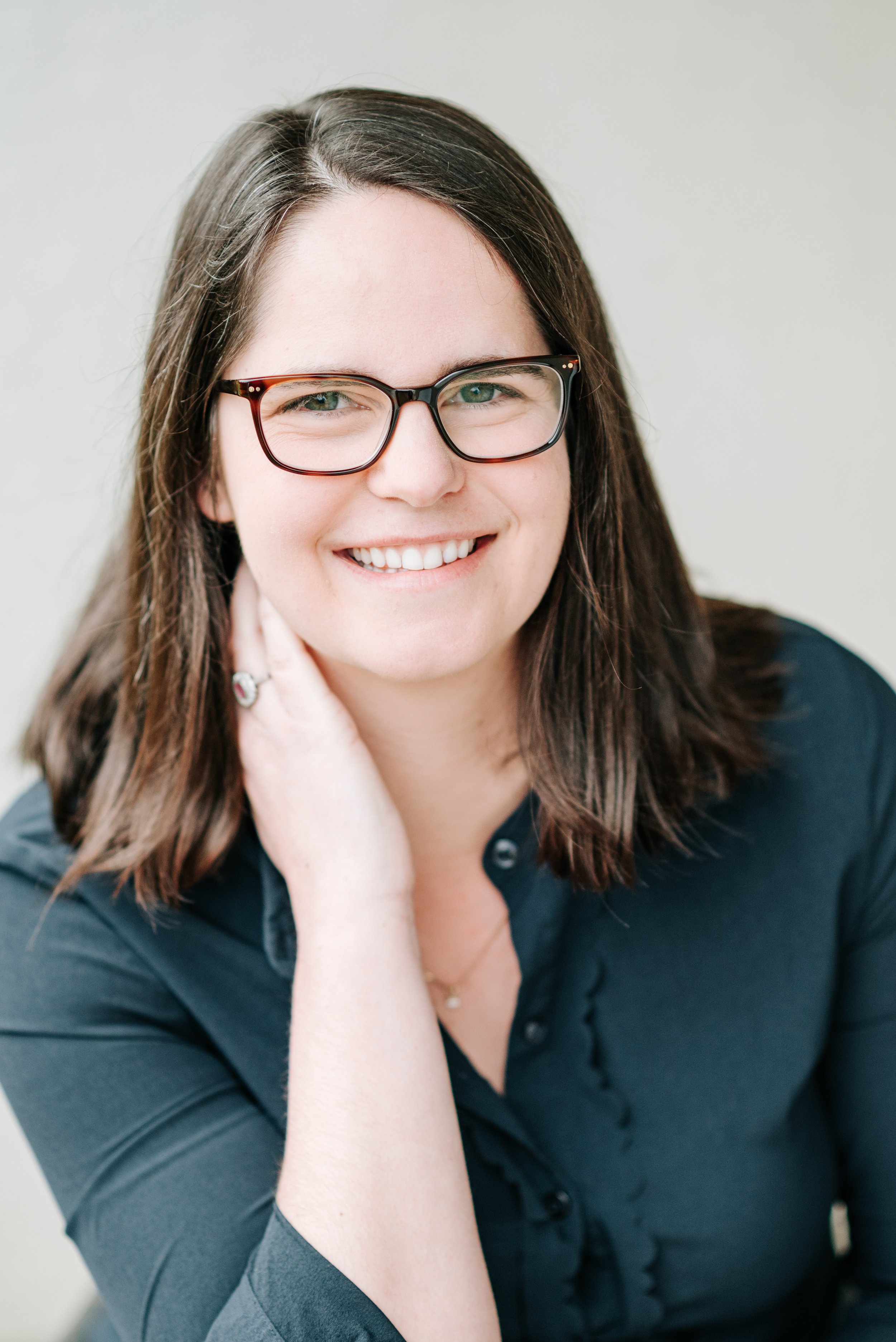 A woman with long brown hair, glasses, and a warm smile, wearing a dark shirt, resting her hand on her neck, against a plain light-colored background.