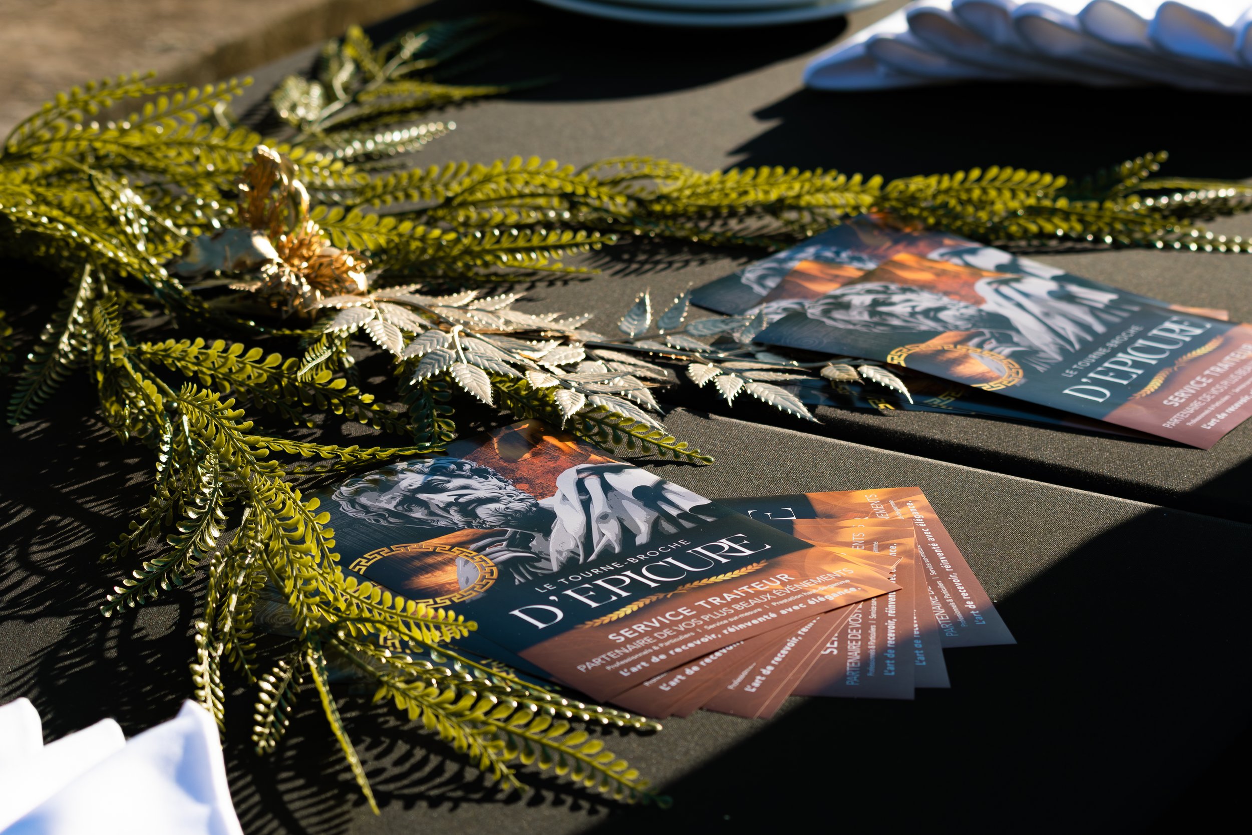 Des brochettes de fleurs vertes et de feuilles, accompagnées de flyers promotionnels pour un événement ou un service décoratif, disposés sur une surface sombre avec de l'éclairage naturel.