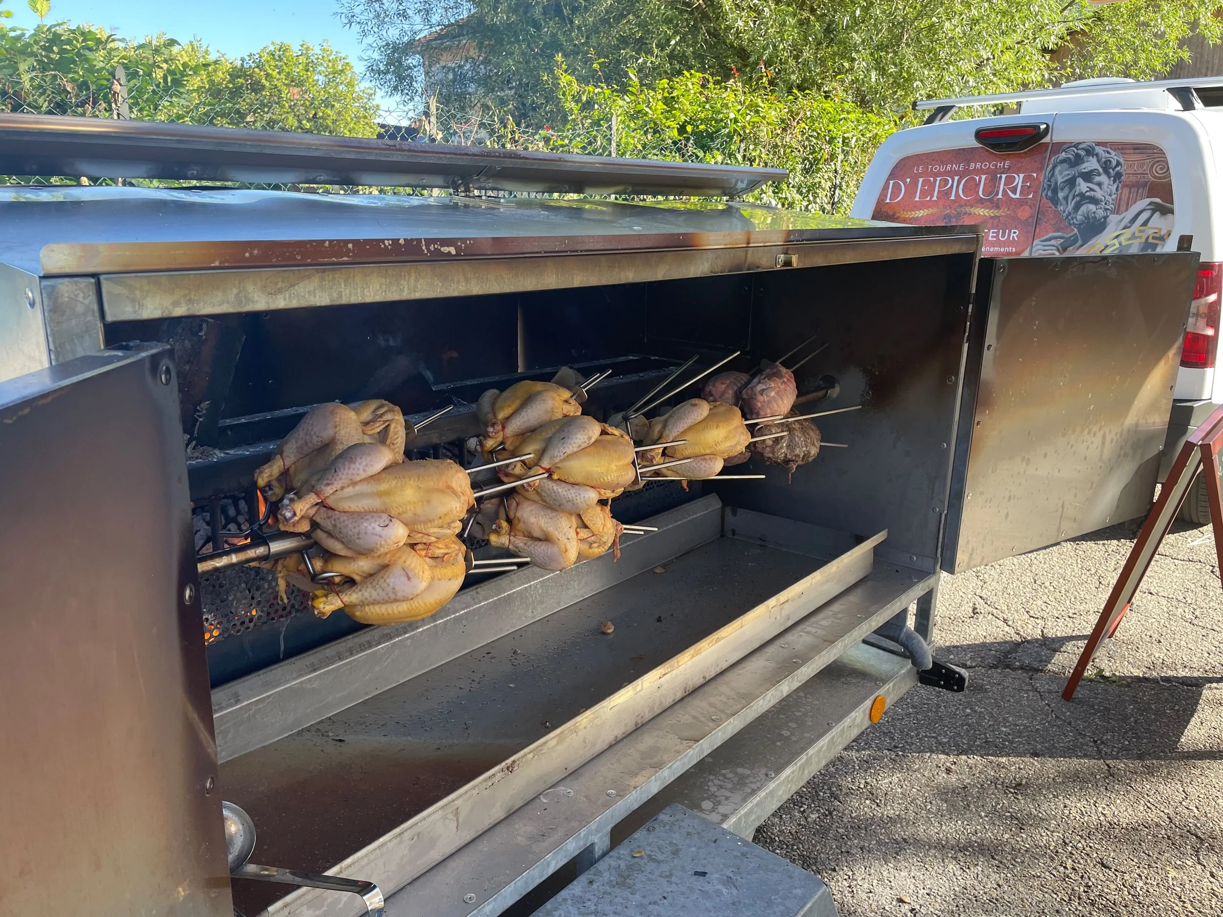 Un barbecue en train de cuire des morceaux de viande et de poulet, avec une remorque de véhicule en arrière-plan décorée d'une image de statue antique et de texte en français.