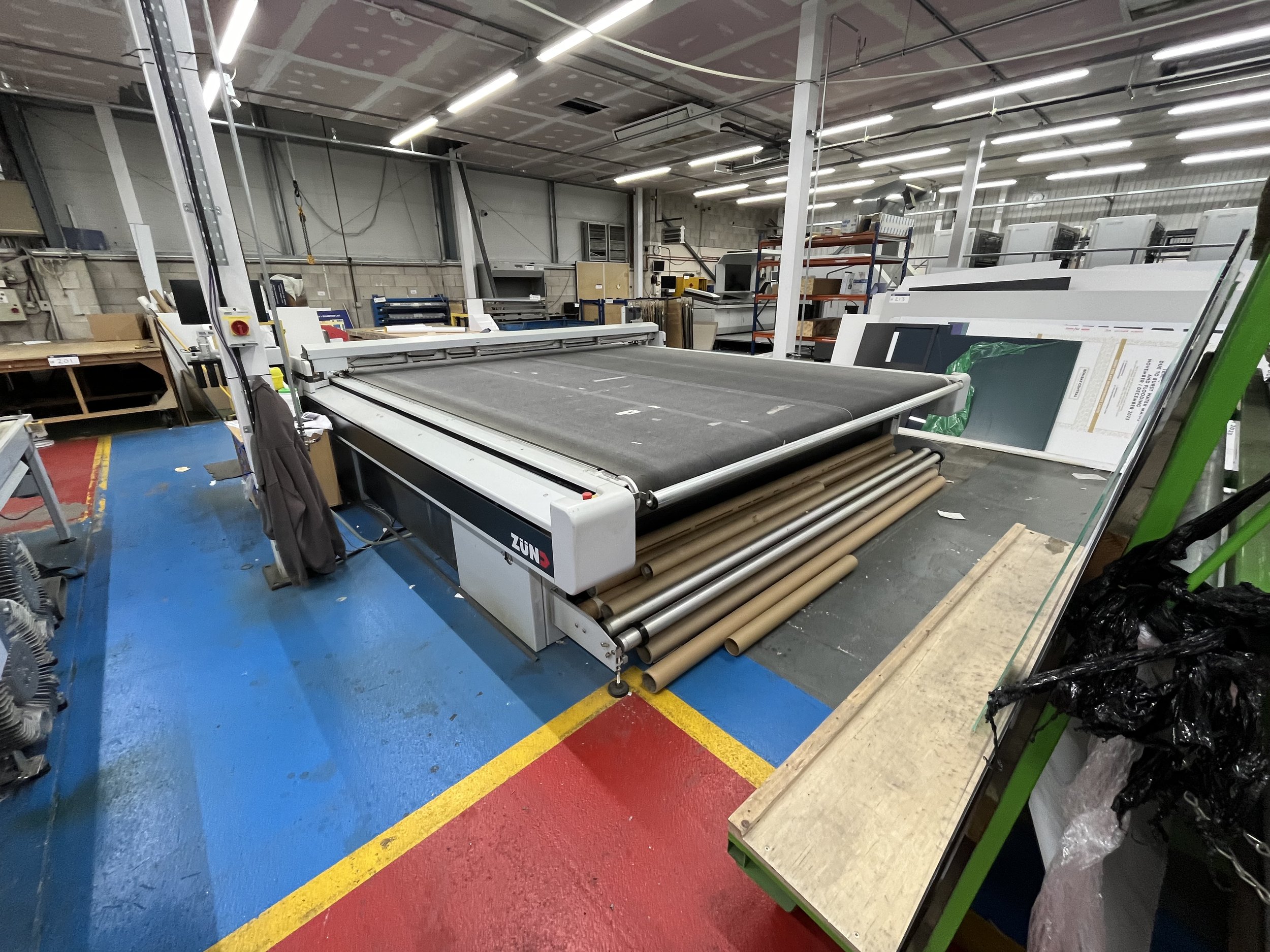 An industrial workspace with a large fabric cutting machine in the foreground, surrounded by rolls of fabric and storage shelves filled with materials and equipment. The area has a concrete wall and ceiling with strip lighting.