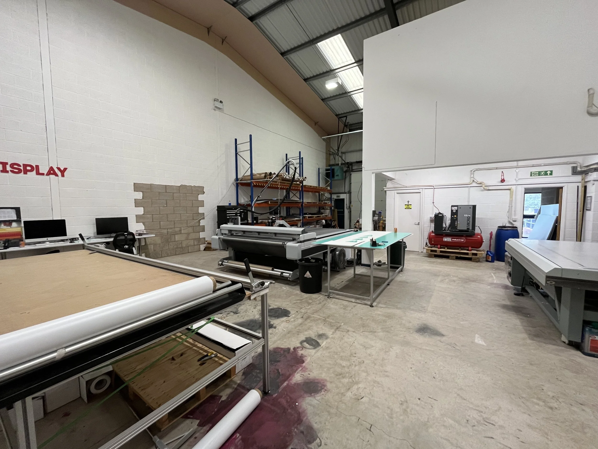 View of an industrial workspace with printing equipment, shelving units, computer monitors, and scattered materials, with a white brick wall and high ceiling.
