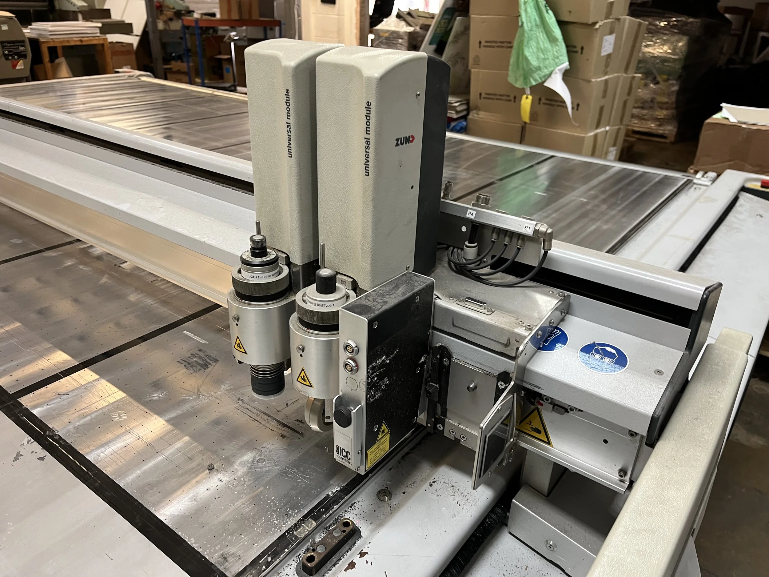 A large industrial machine with metal components and wiring, situated in a workshop environment with cardboard boxes and shelves in the background.