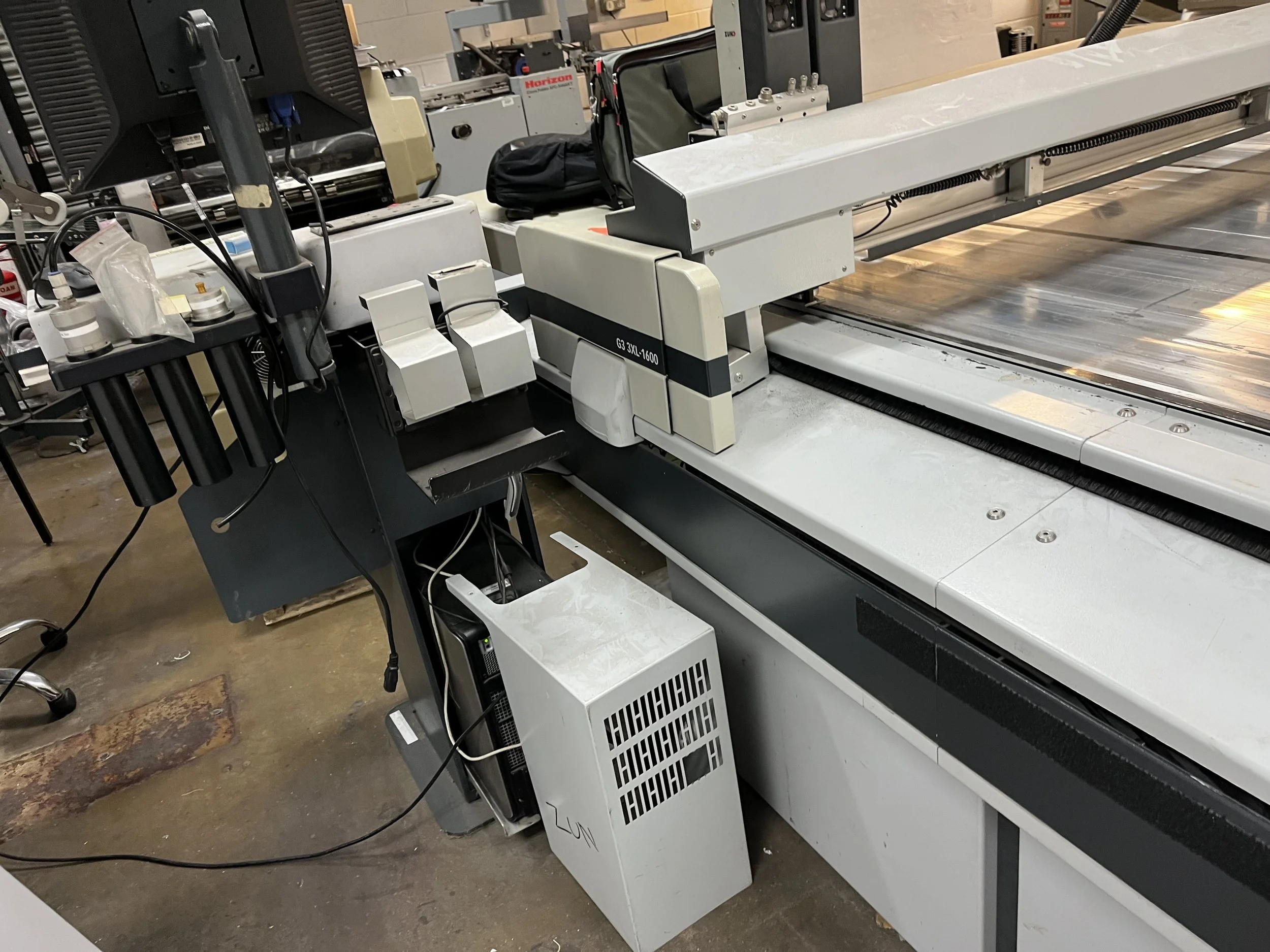 A large industrial printing or printing-like machine with a metallic and white structure, featuring various belts, rollers, and electronic components, situated in a warehouse or workshop setting.