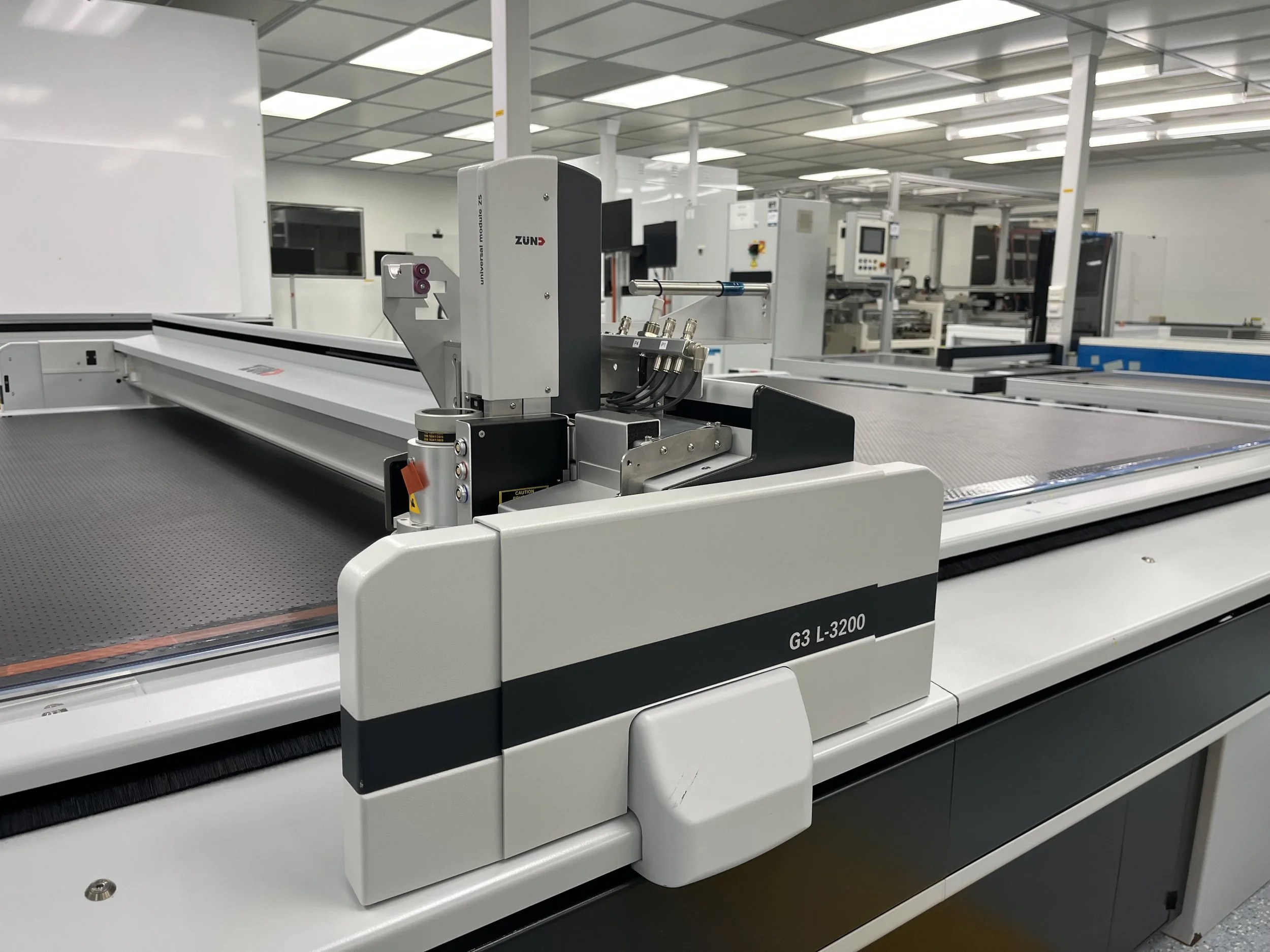 Inside a modern factory or laboratory with automated machinery. There is a large white manufacturing or testing system with label G3 L-3200 and various components, situated on a gray textured surface, and additional equipment and screens are visible 