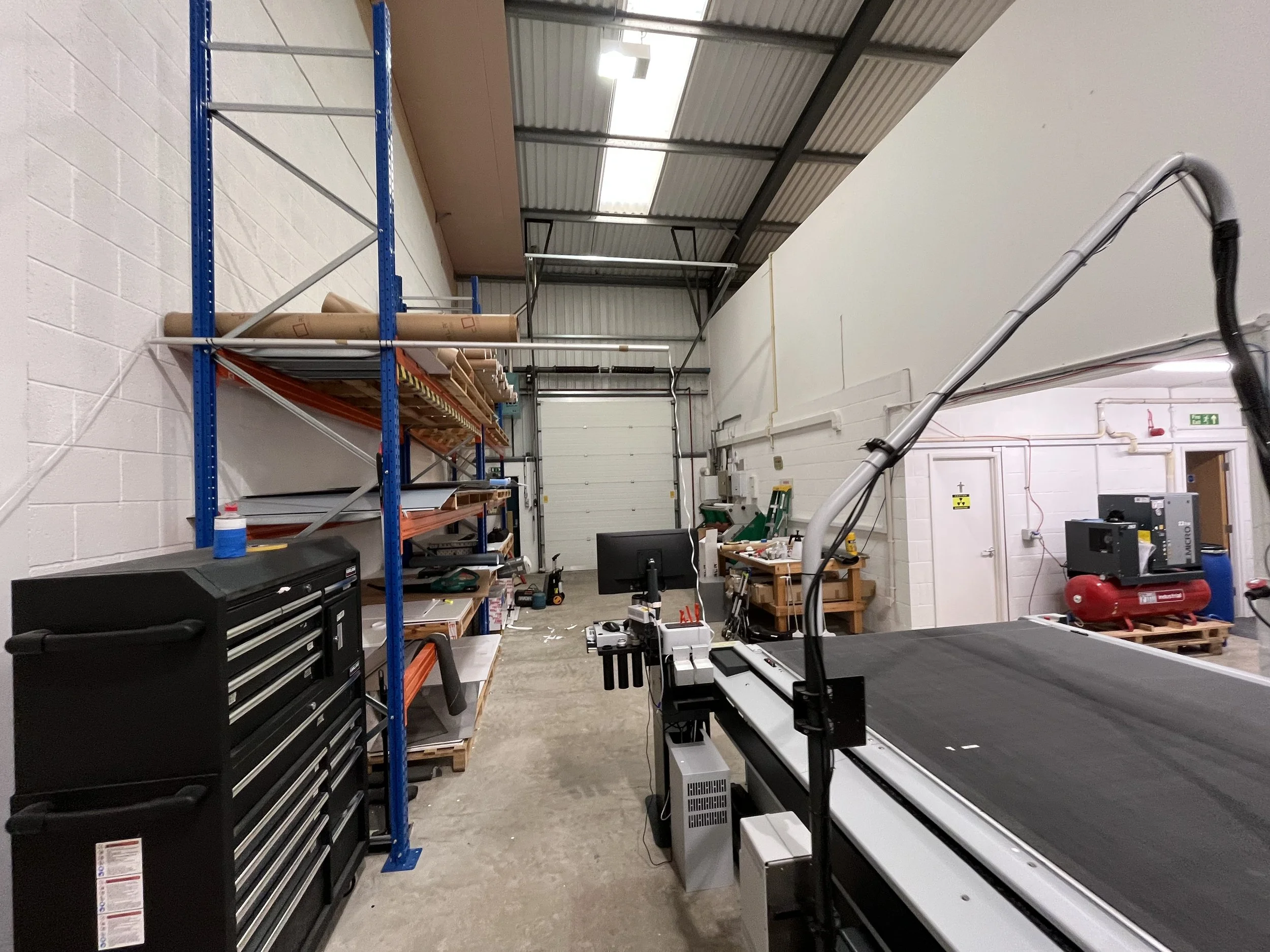 A workshop or storage area with a metal shelving unit holding cardboard and wooden materials, a work table with tools, a black tool chest, and a large machine or work station with a sliding rail and a vacuum hose, in a space with white brick walls an