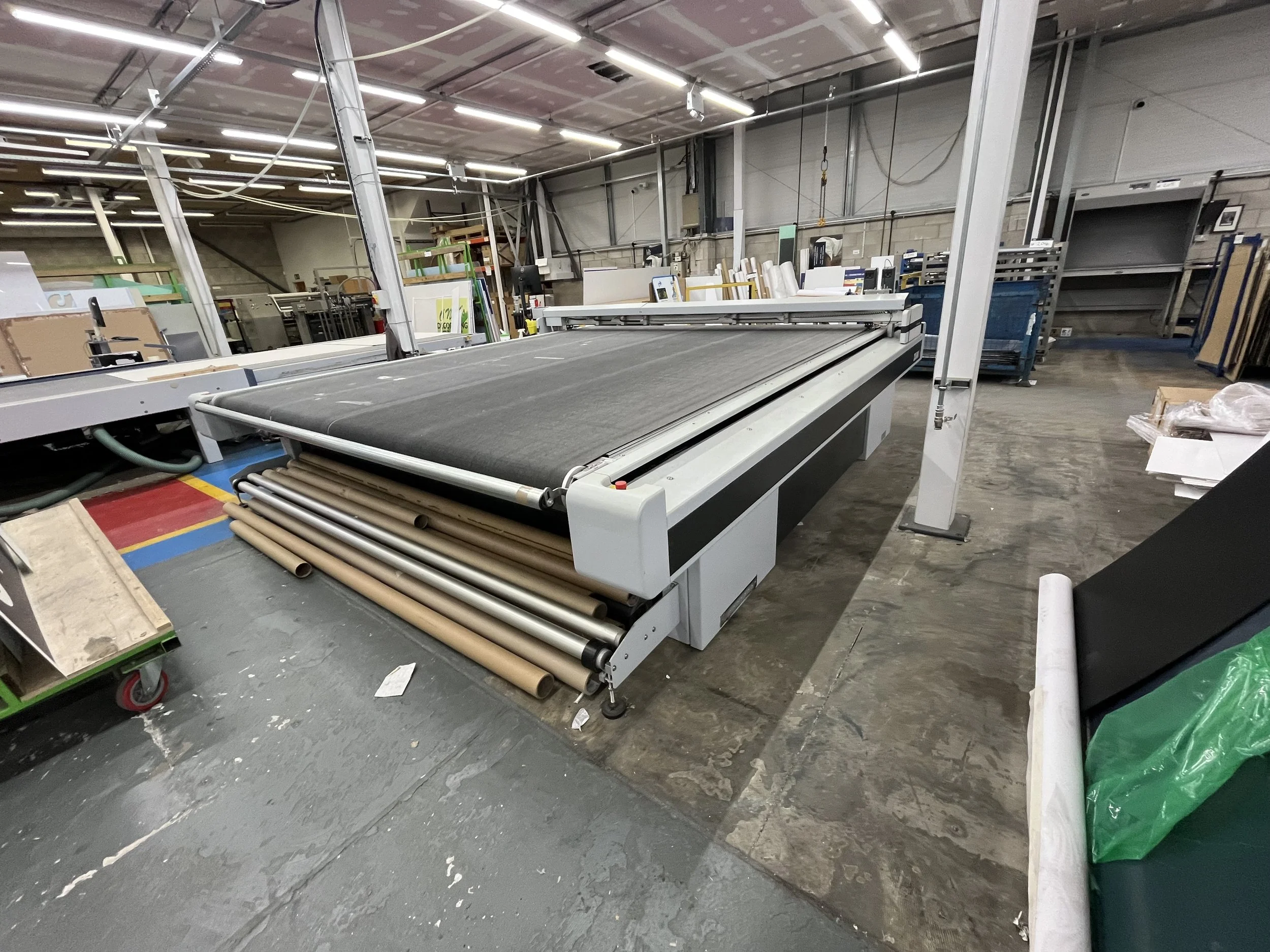Empty industrial workshop with a large machine, rolls of paper or material underneath, surrounded by shelves, boxes, and various equipment, with high ceilings and bright fluorescent lighting.