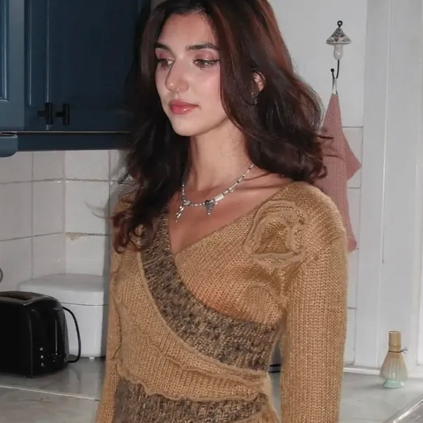 A woman with long, dark hair and light skin wearing a tan and brown sweater, standing in a kitchen with white tiled walls and blue cabinets.