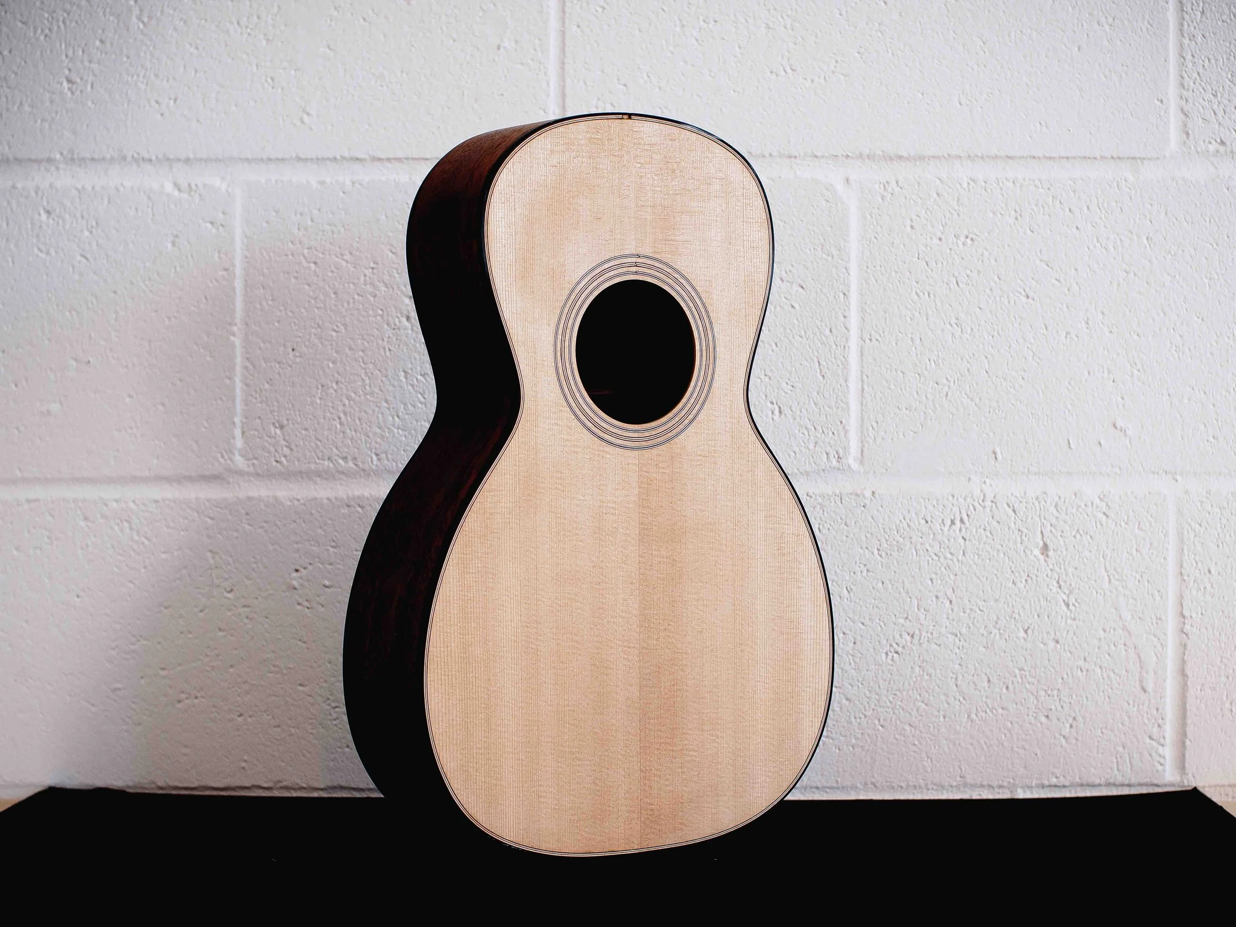 An acoustic guitar, placed against a white brick wall, with shadows cast on a black surface.