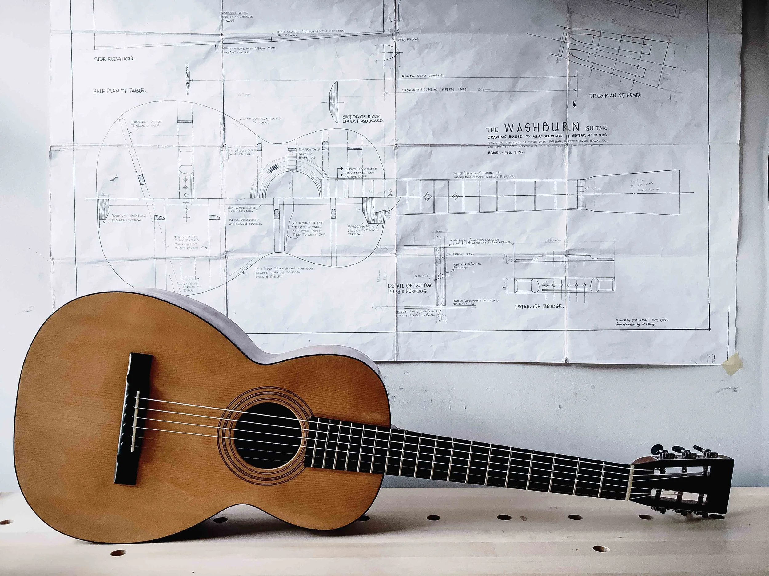 An acoustic guitar resting against a large detailed blueprint or technical drawing of a woodworking project on a wall.