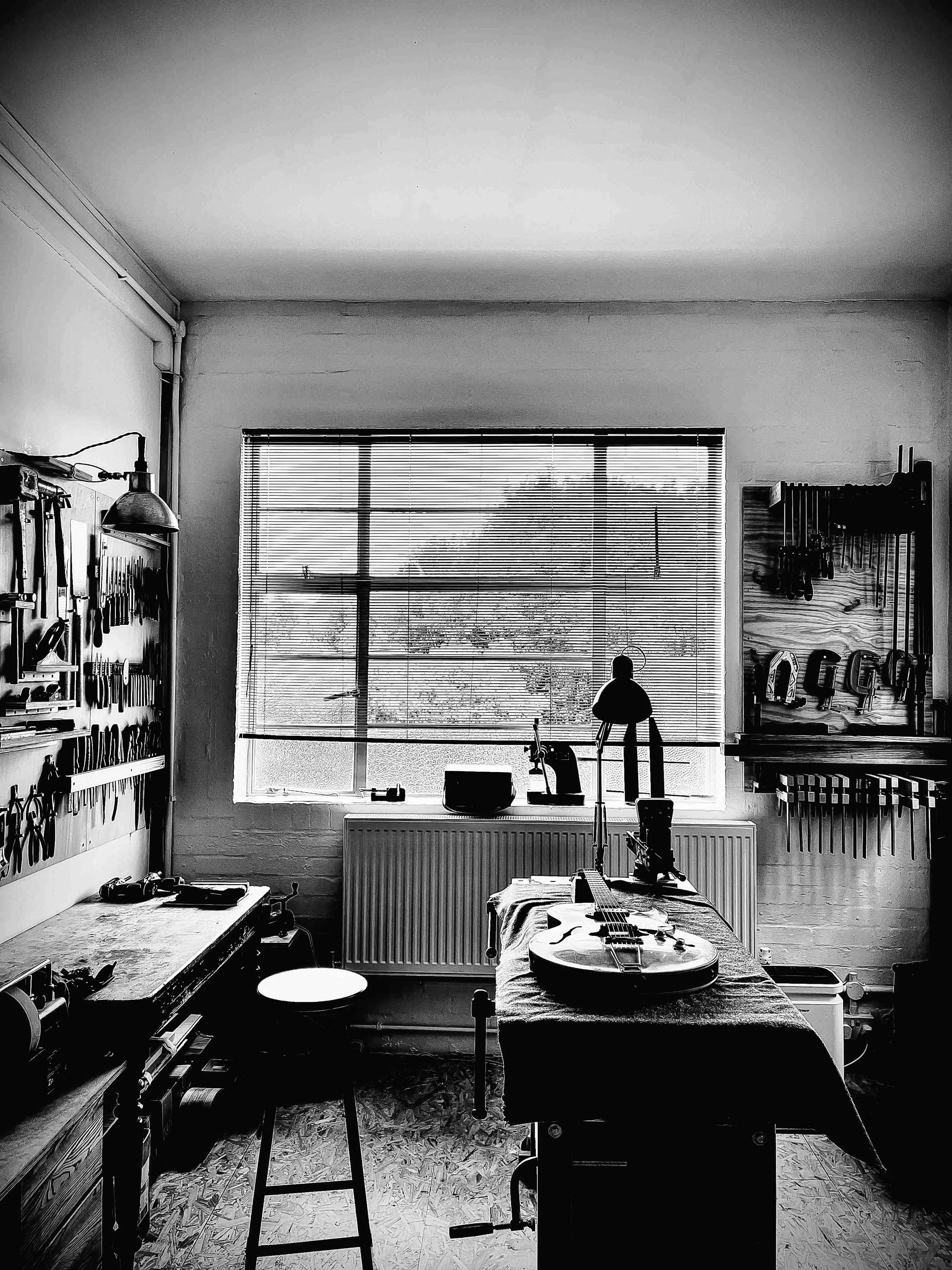 A black-and-white photo of a workshop with an electric guitar on a table, a workbench with tools, a window with blinds, and various musical and workshop equipment.