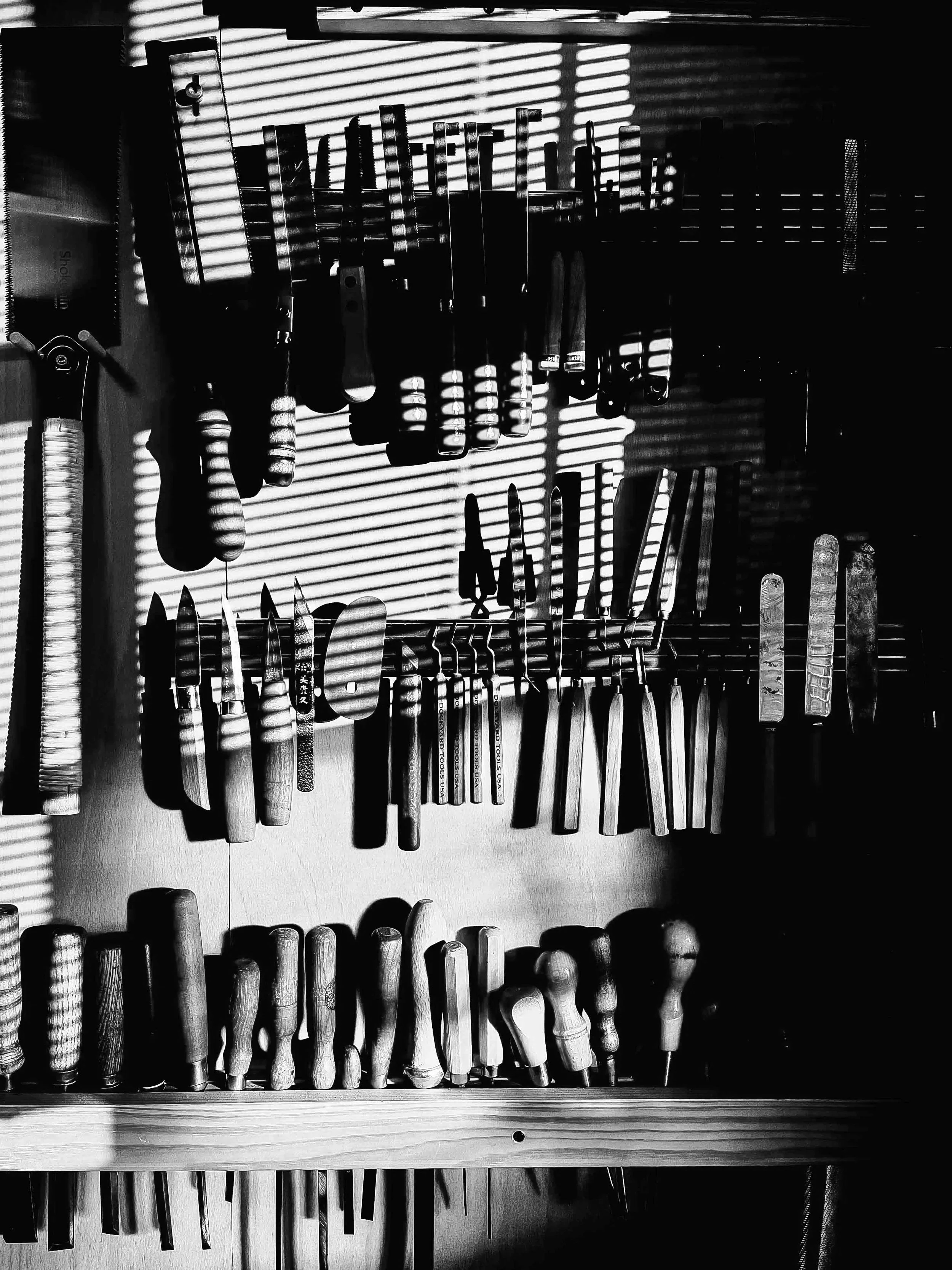 Collection of various knives and woodworking tools hanging on a wall with sunlight casting striped shadows.