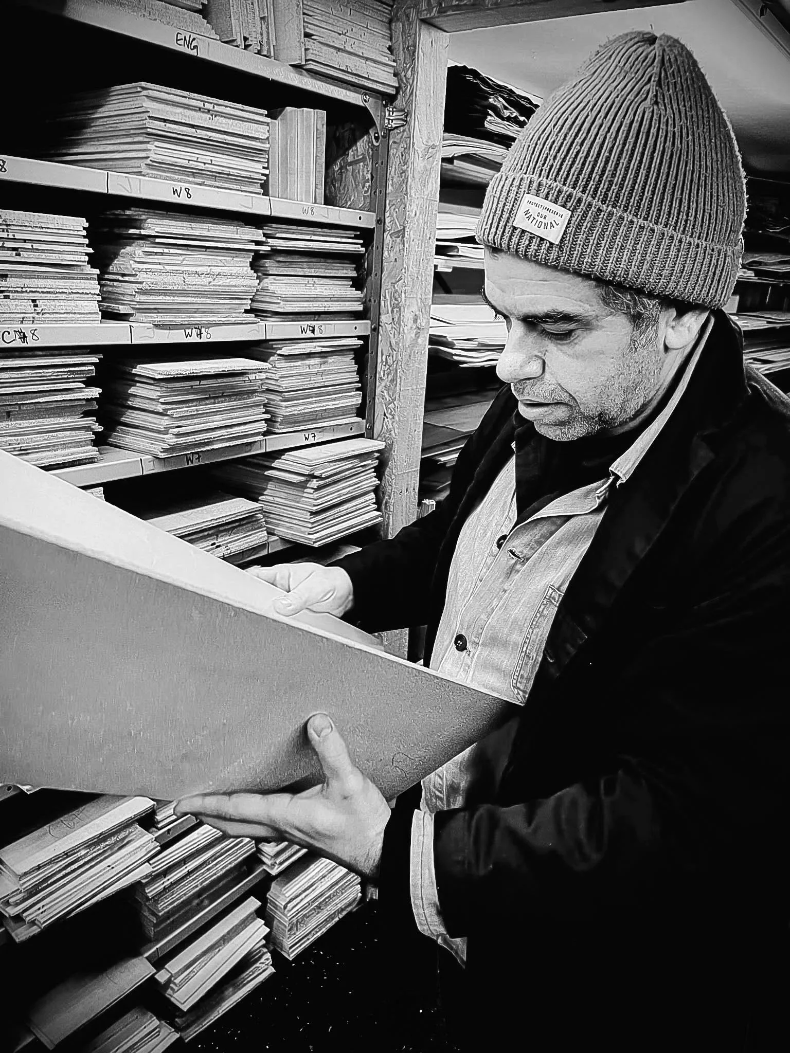 A person wearing a beanie and coat, selecting acoustic guitar tops from shelves labeled with categories, in a wood merchants.