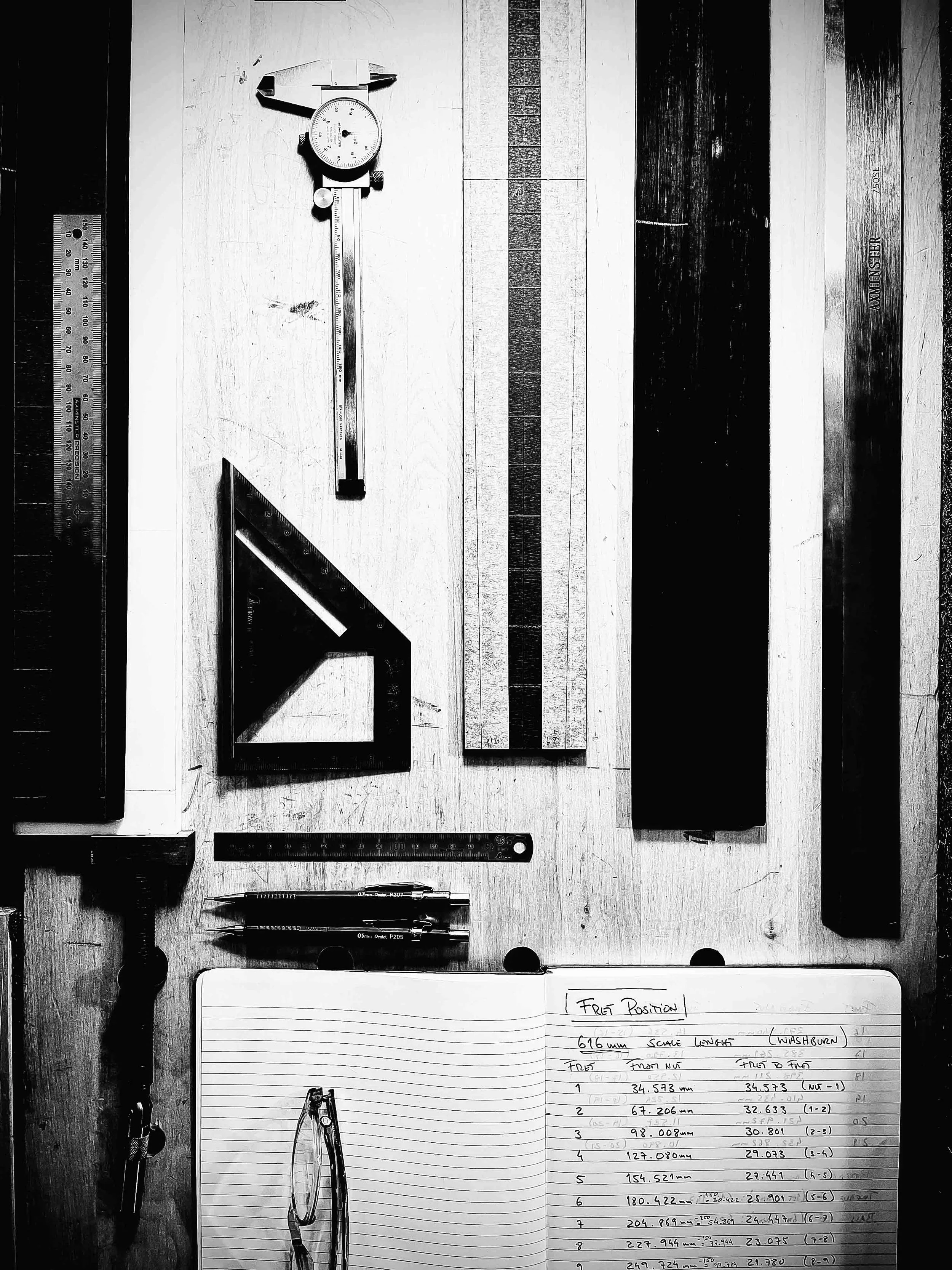 A black and white photo of a work desk with a digital caliper, a metal ruler, a pair of compasses, and a notebook with handwritten notes and measurements on lined paper. The notebook page has the words 'Fret Position' and a list with dimensions.