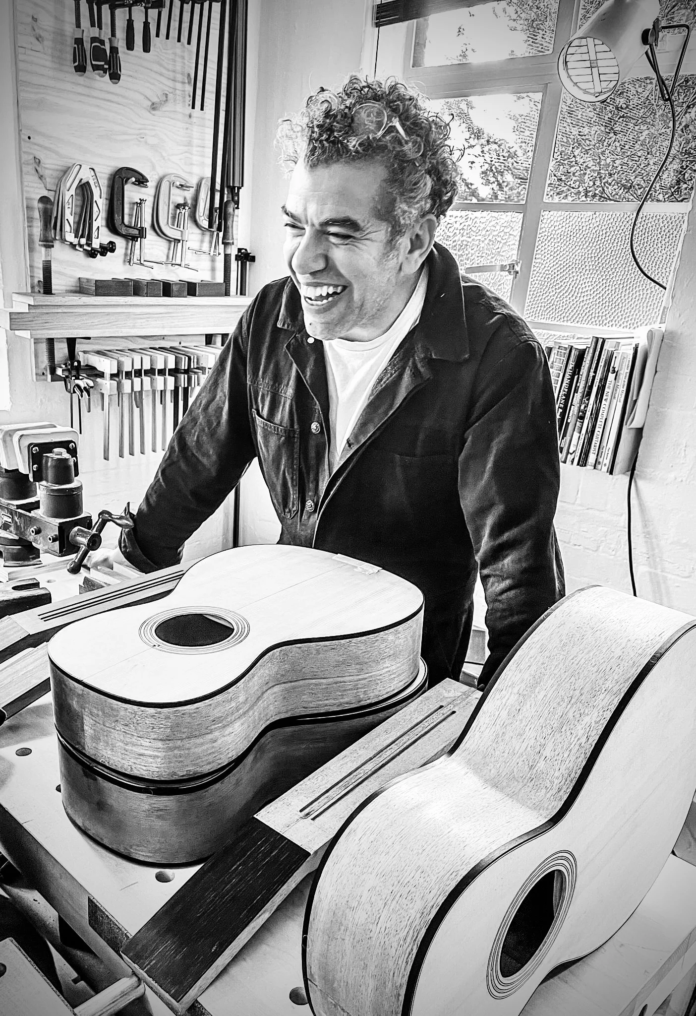 A man with curly hair smiling while working on a guitar in a workshop with woodworking tools on the wall. There are large windows behind him and the workshop contains various woodworking equipment and books.
