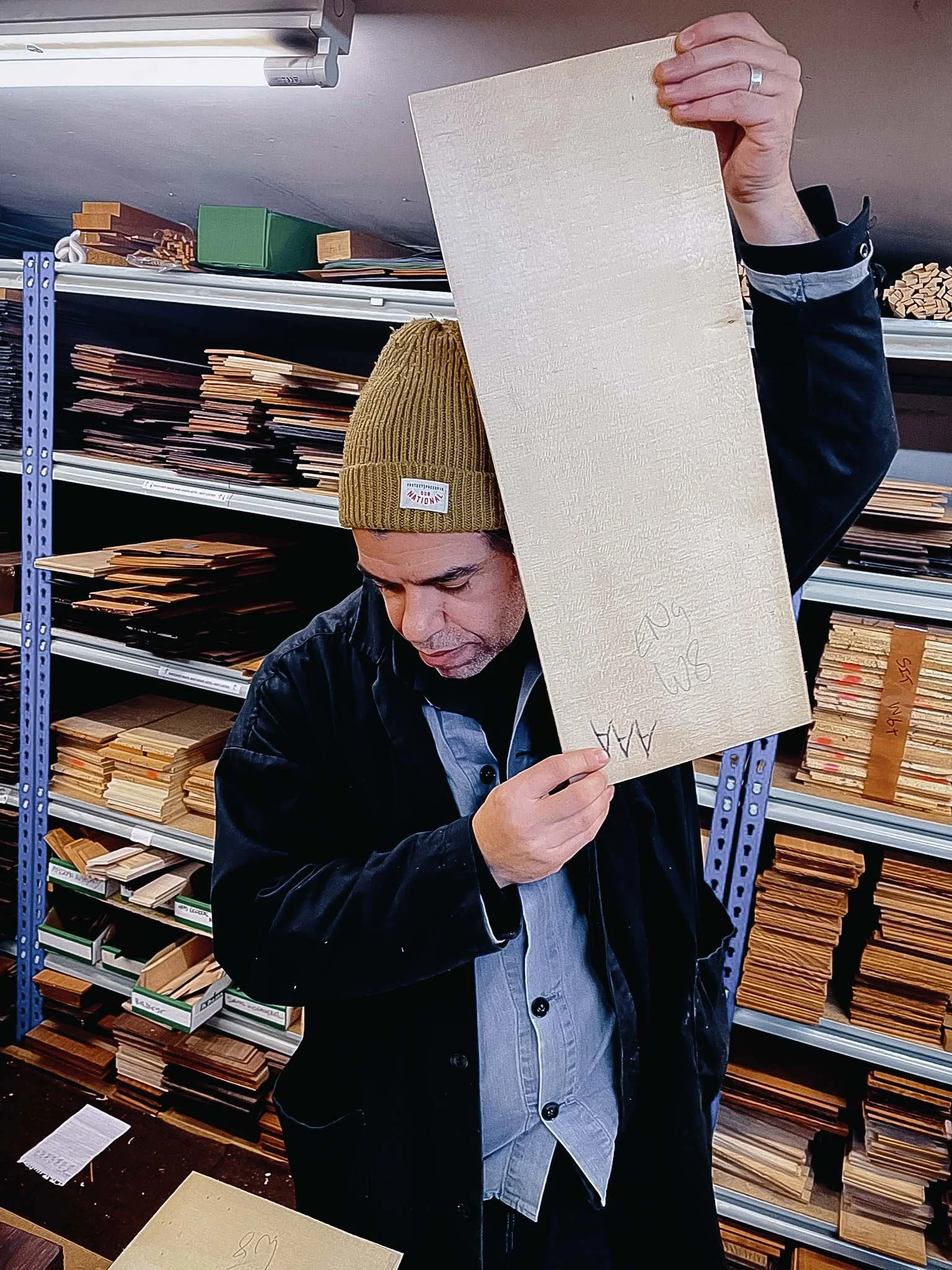 Man wearing a brown knit beanie and black jacket holding a guitar soundboard in a store with shelves of wood samples in the background.