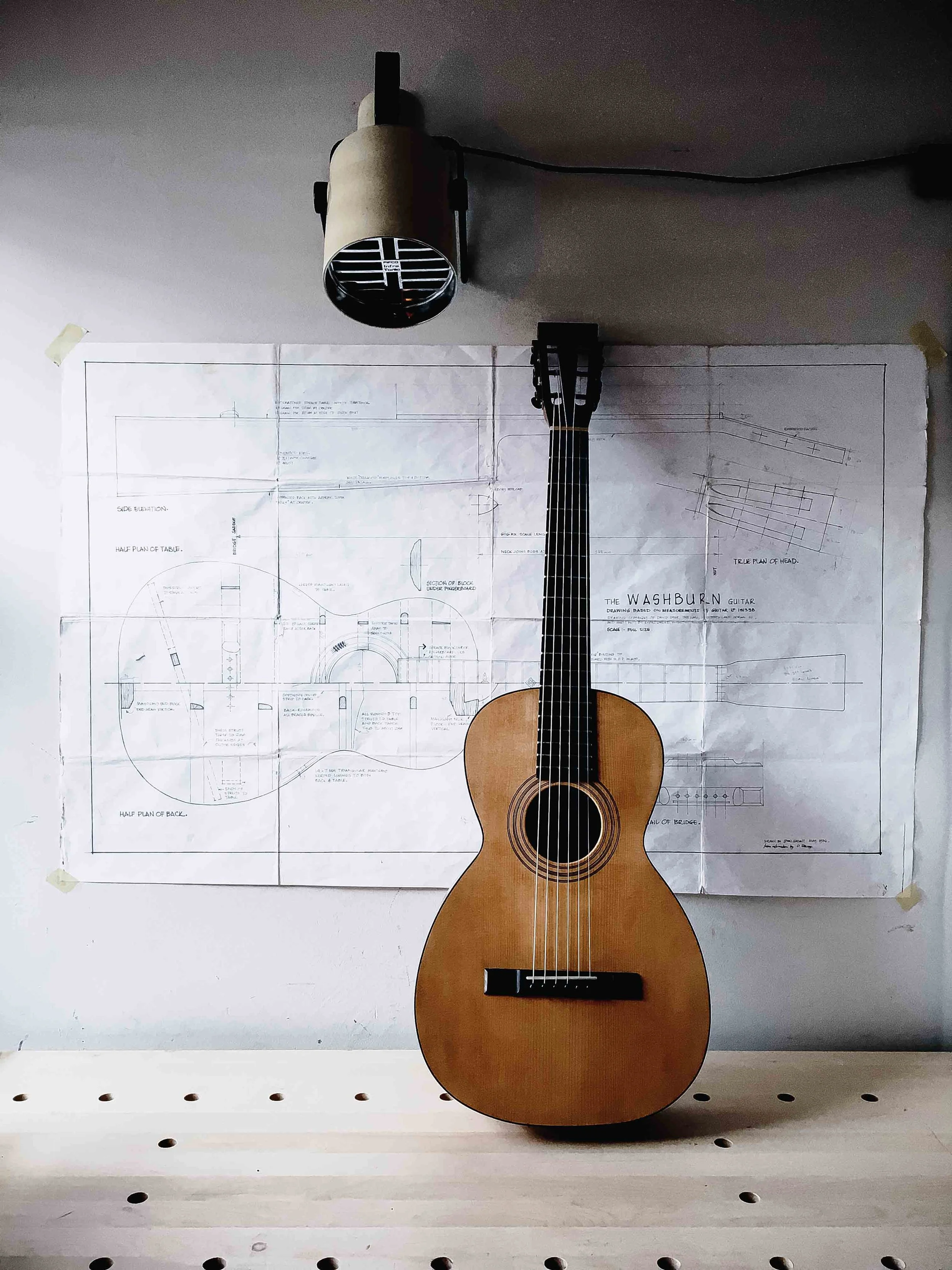 An electric guitar is mounted on a wall with architectural blueprints behind it. The blueprints are taped to the wall and feature detailed drawings and notes for building a guitar, titled 'The Washburn Guitar'. The guitar has a natural wood finish, six strings, and a black bridge and tuning pegs.