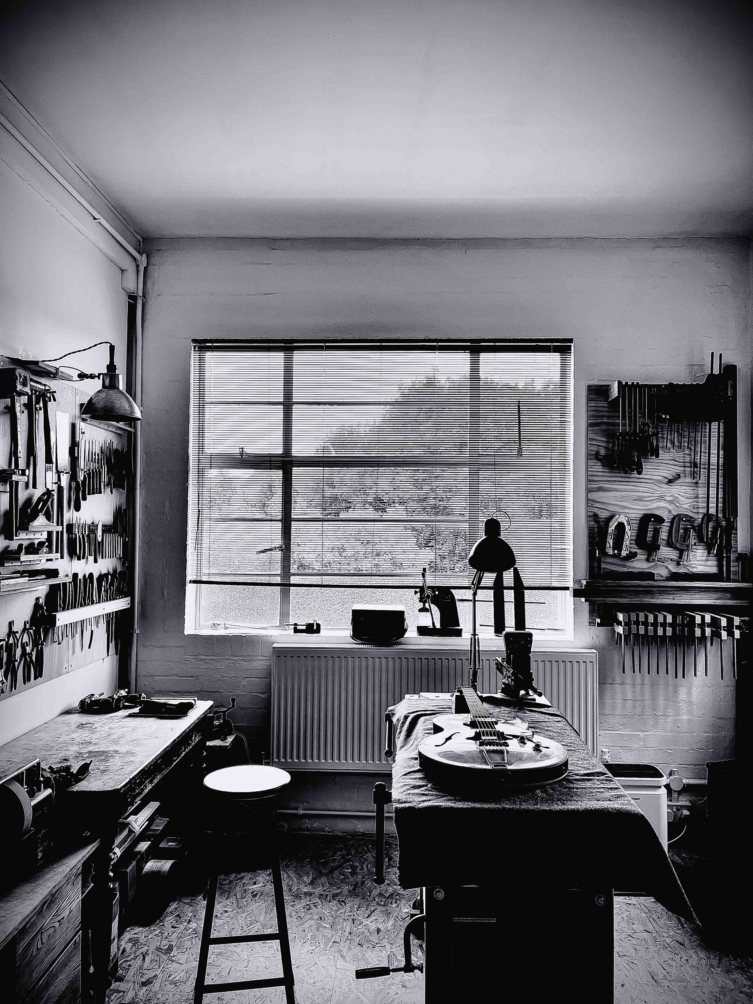 A black and white photo of a room with a large window with blinds, a workbench with tools on the left wall, a guitar on a table in the foreground, and various tools hanging on the right wall. There is a lamp illuminating the workspace.