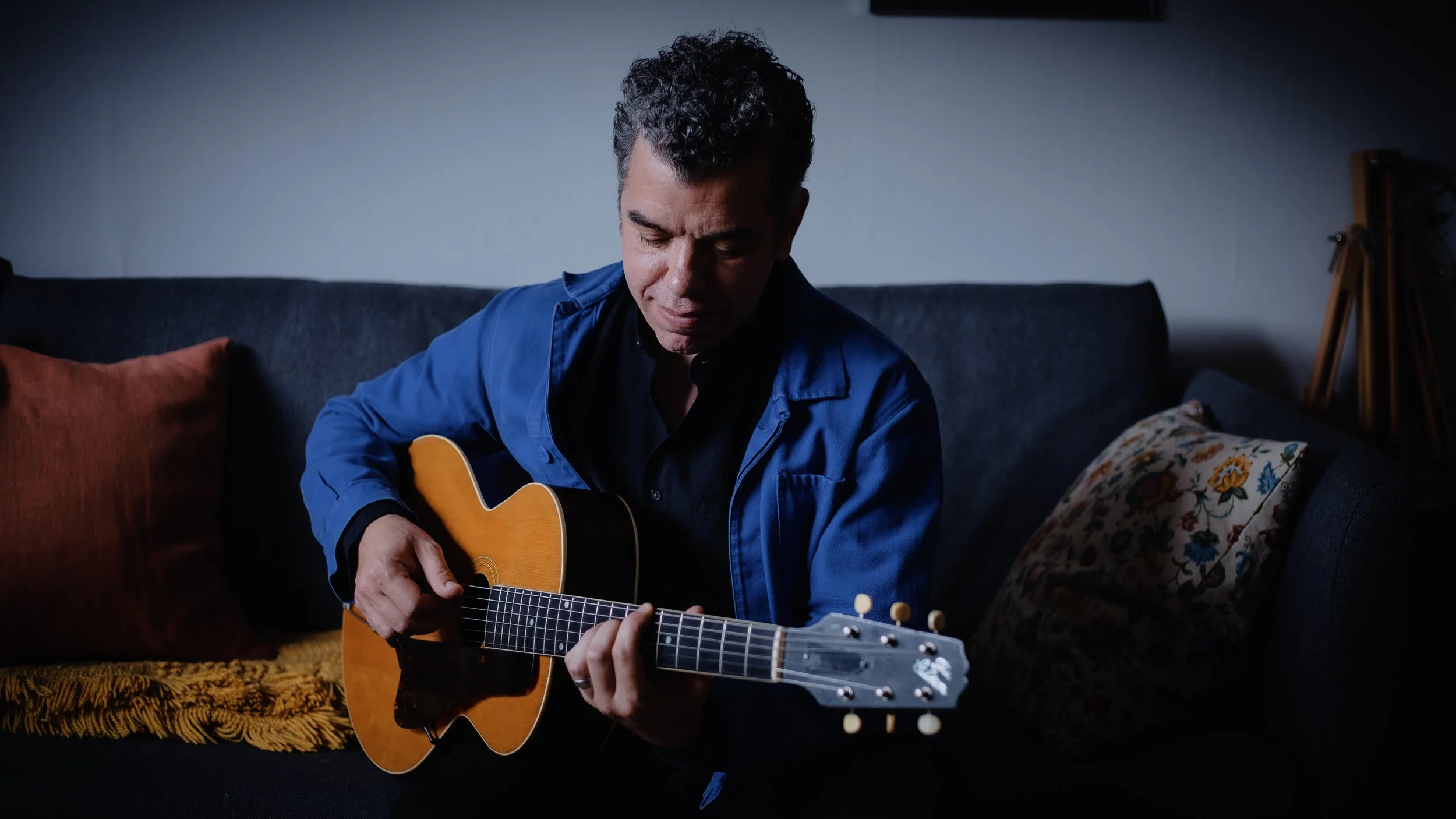 A man with curly dark hair wearing a blue jacket and black shirt playing an acoustic guitar while sitting on a dark-colored sofa decorated with colorful pillows in a dimly lit room.