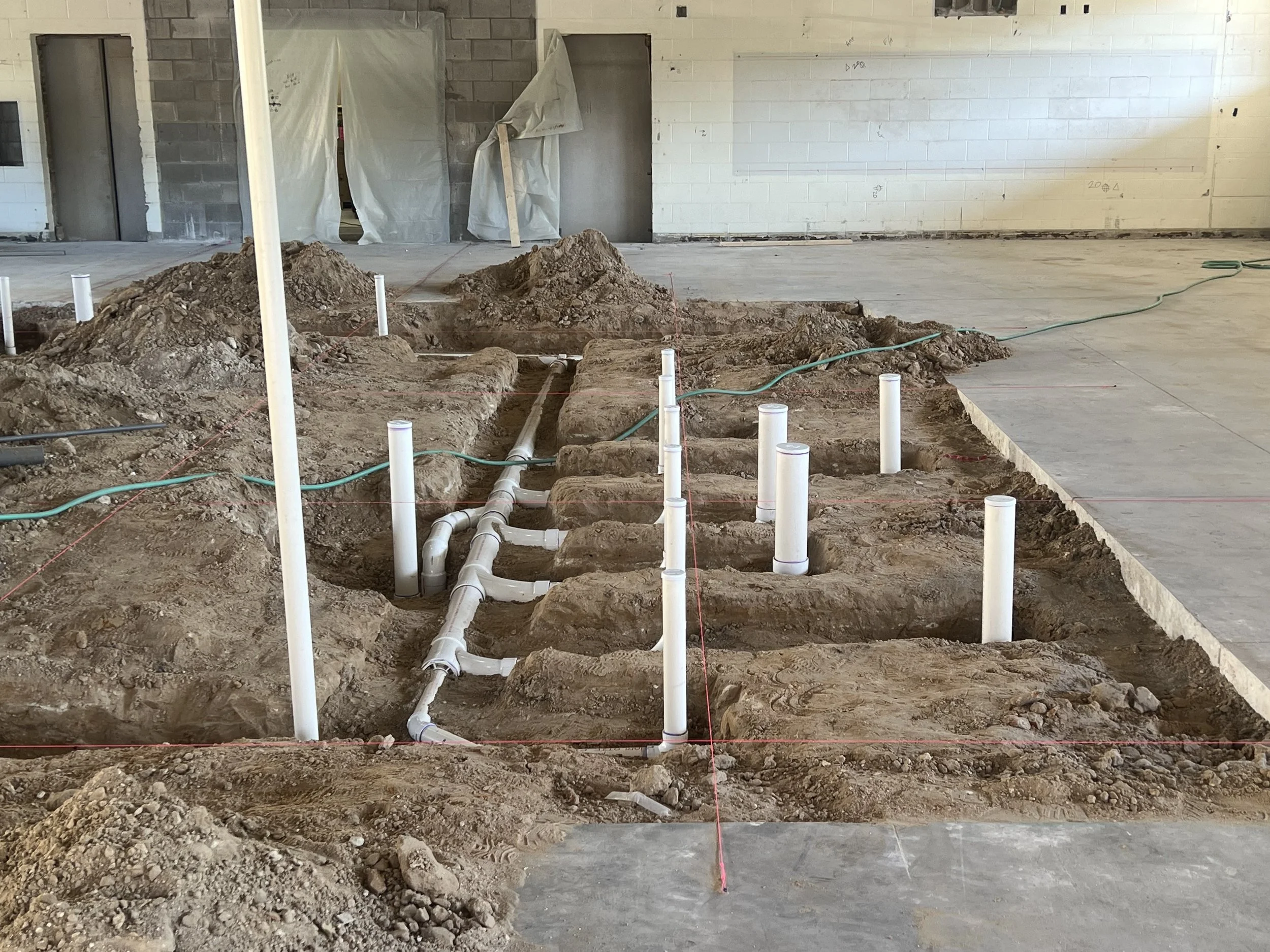 Construction site with exposed pipes and dug trenches in the floor, with drywall and construction materials on the walls in the background.
