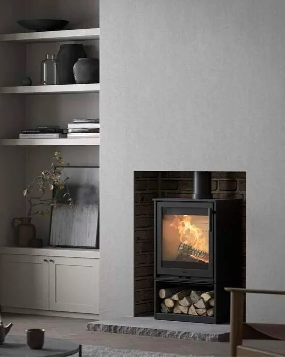 A modern living room corner with a wood-burning stove in a brick-lined niche, fire visible inside, and a built-in firewood storage below. To the left, there is a built-in white shelf with decorative vases, books, and a dried branch in a vase, all against a light gray wall.