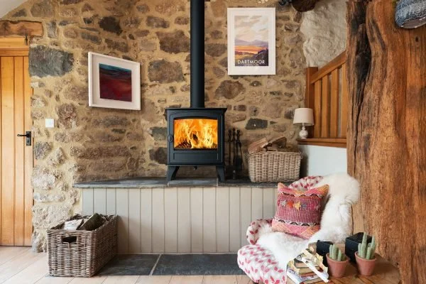 Living room with stone wall, a wood-burning stove with fire, framed artwork on the wall, a wicker basket, a cozy chair with patterned blanket and pillow, and small potted plants.