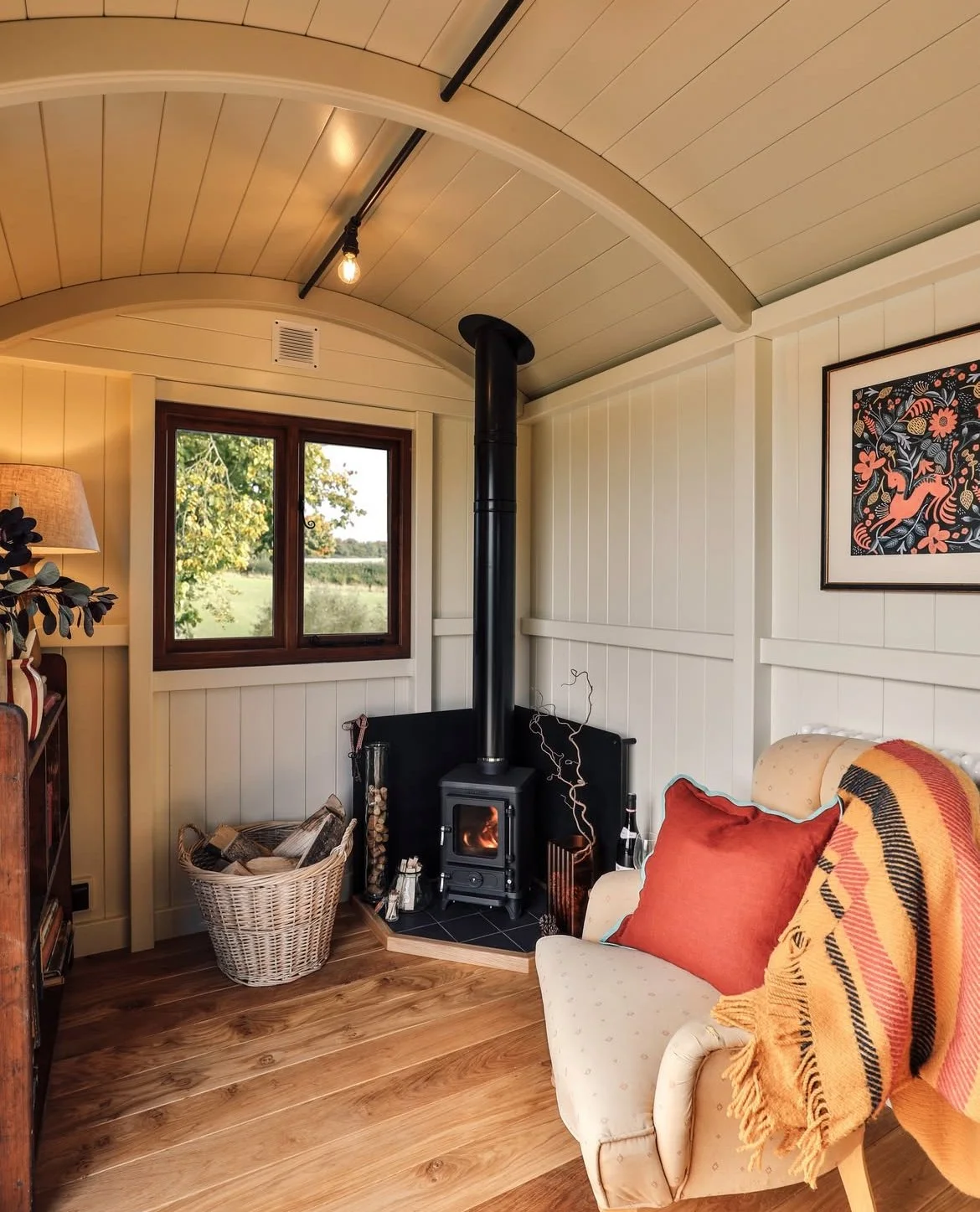 Living room with wood-paneled walls, small wood stove, basket of firewood, window showing green trees, armchair with orange pillow and a blanket, framed artwork on wall, wooden floor, and ceiling with exposed bulb light fixture.
