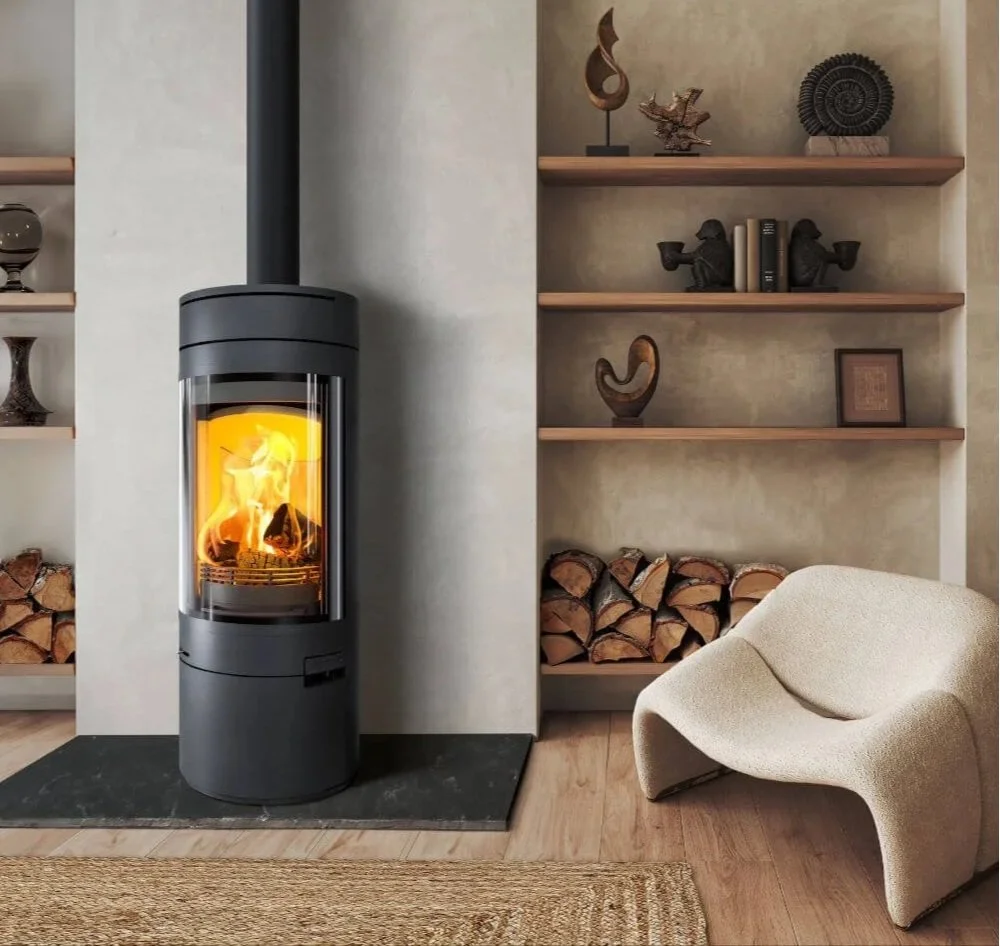 Modern living room with a black wood-burning stove on a stone hearth, surrounded by built-in shelves with decorative items and stacked firewood, and a cozy beige armchair.
