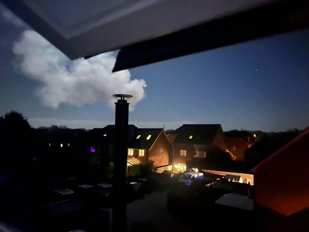 Nighttime view from a window showing houses with illuminated windows, parked cars, a chimney emitting smoke, and a cloudy sky with a visible star.