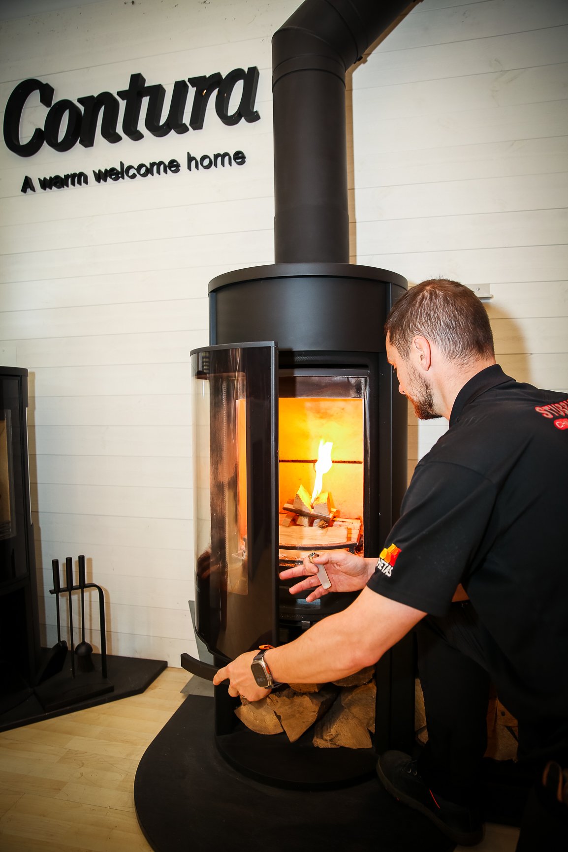 A man is adjusting a fire-starting device in front of a wood stove at Contura, a home decor store. The store wall has a sign that reads, "Contura: A warm welcome home."
