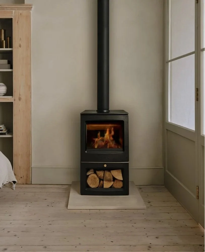 Black wood-burning stove with fire inside and stacked firewood in a compartment below, located in a cozy room with light wooden floors, a beige wall, a window, and a bookshelf nearby.