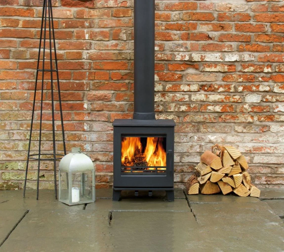A small black wood stove with a fire inside, sitting on a concrete patio against a red brick wall. To the left of the stove, there is a metallic lantern with a handle and a candle inside. To the right, there is a pile of chopped firewood stacked agai