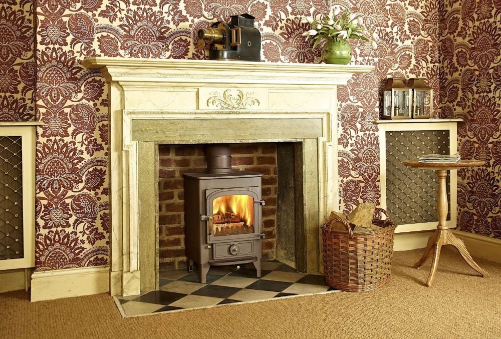 A cozy living room with a fireplace, a wood stove with a fire, a basket of firewood, patterned wallpaper, a small wooden side table, a decorative green vase with flowers, and various small decorative objects on the mantle and shelves.