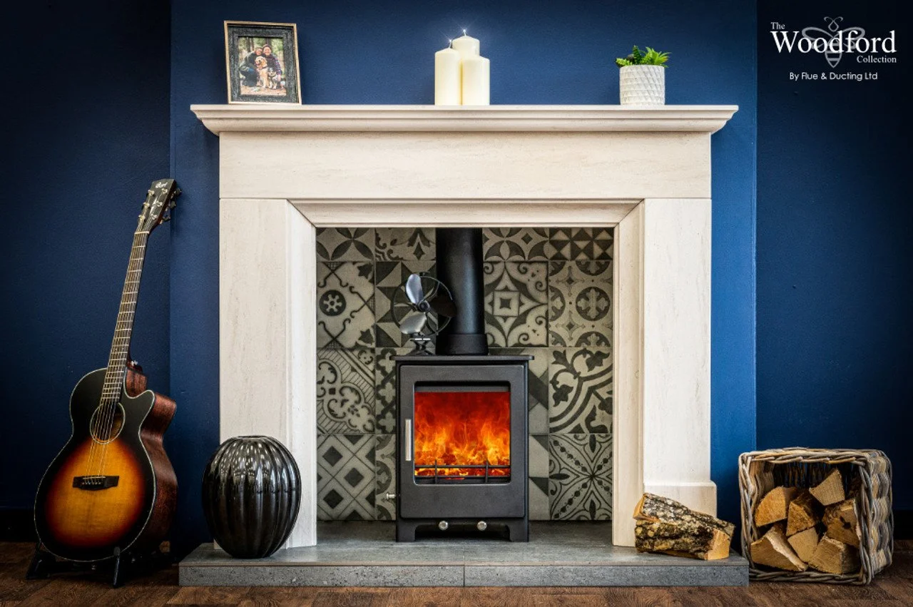 A fireplace with a wood-burning stove in a room with navy blue walls. On the mantel, there is a framed family photo, two candles, and a potted plant. To the left of the fireplace, an acoustic guitar leans against the wall, and next to it is a black d